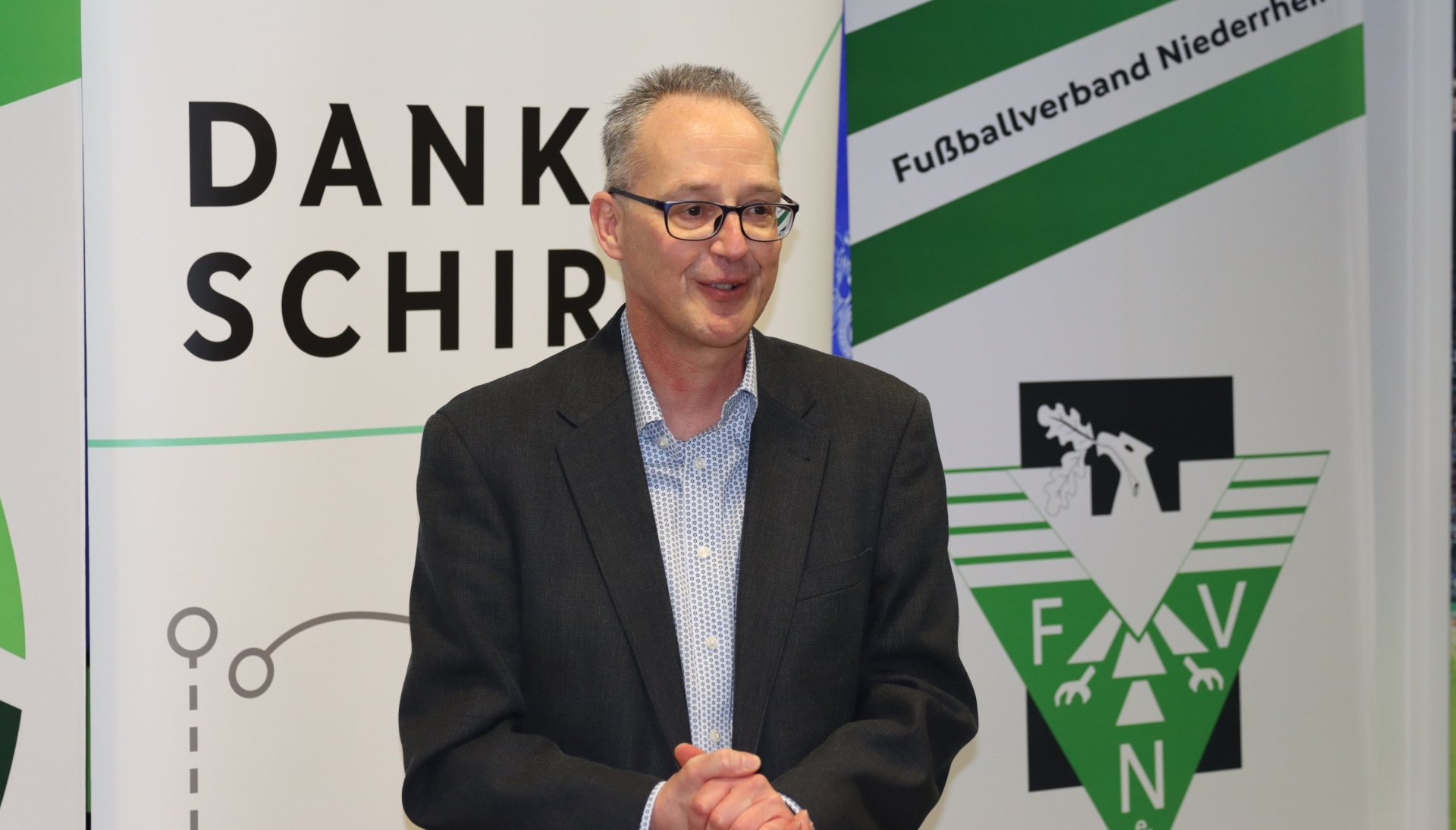 [{"clang_id":1,"value":"Mann mit Brille im Anzug vor Hintergrund mit Schriftzug \"DANK SCHIR\" und Logo \"FVN Fußballverband Niederrhein\"."}]