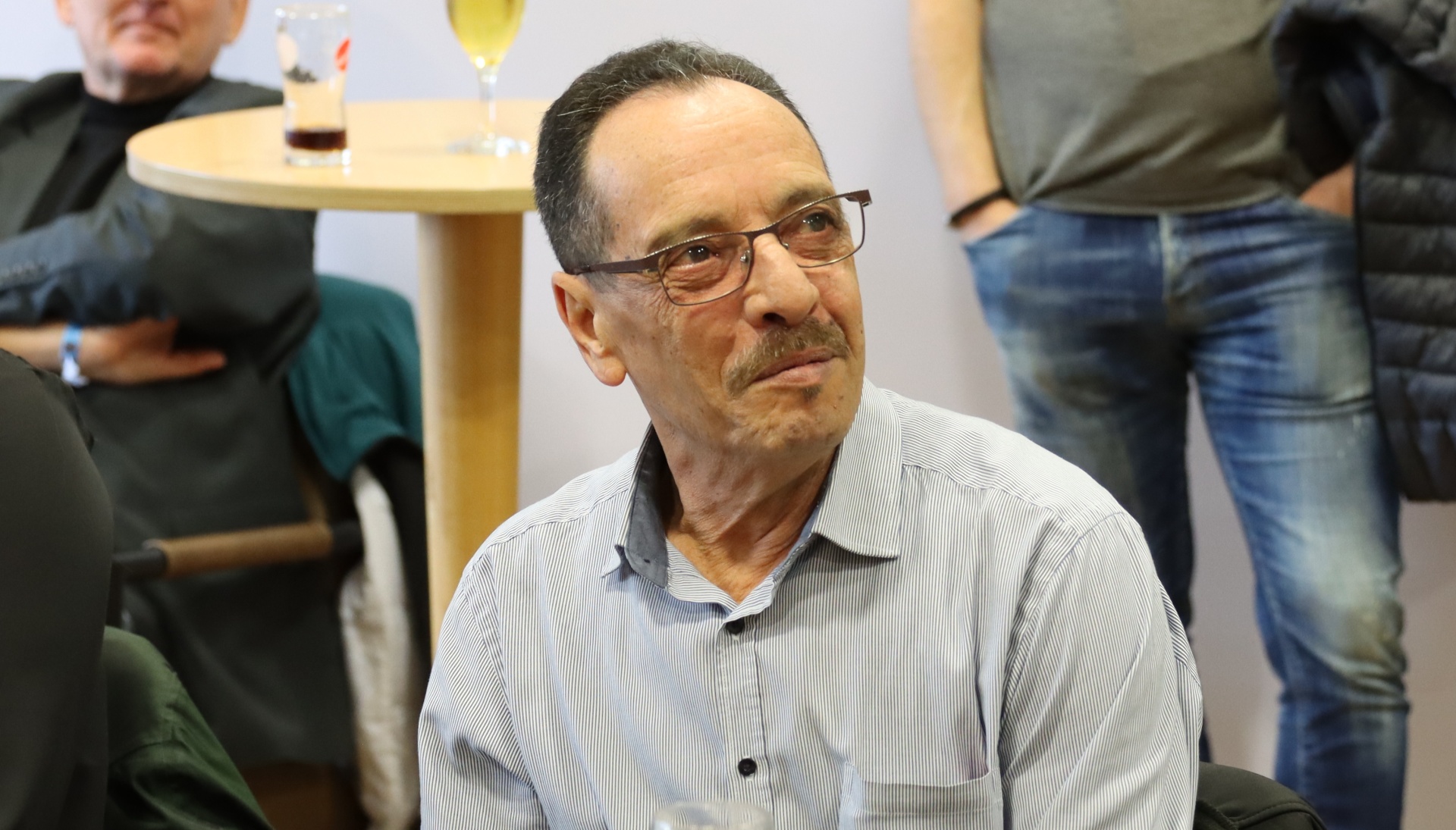 [{"clang_id":1,"value":"Mann mit Brille und gestreiftem Hemd sitzt, blickt zur Seite. Im Hintergrund Personen und ein Tisch mit Getränken."}]