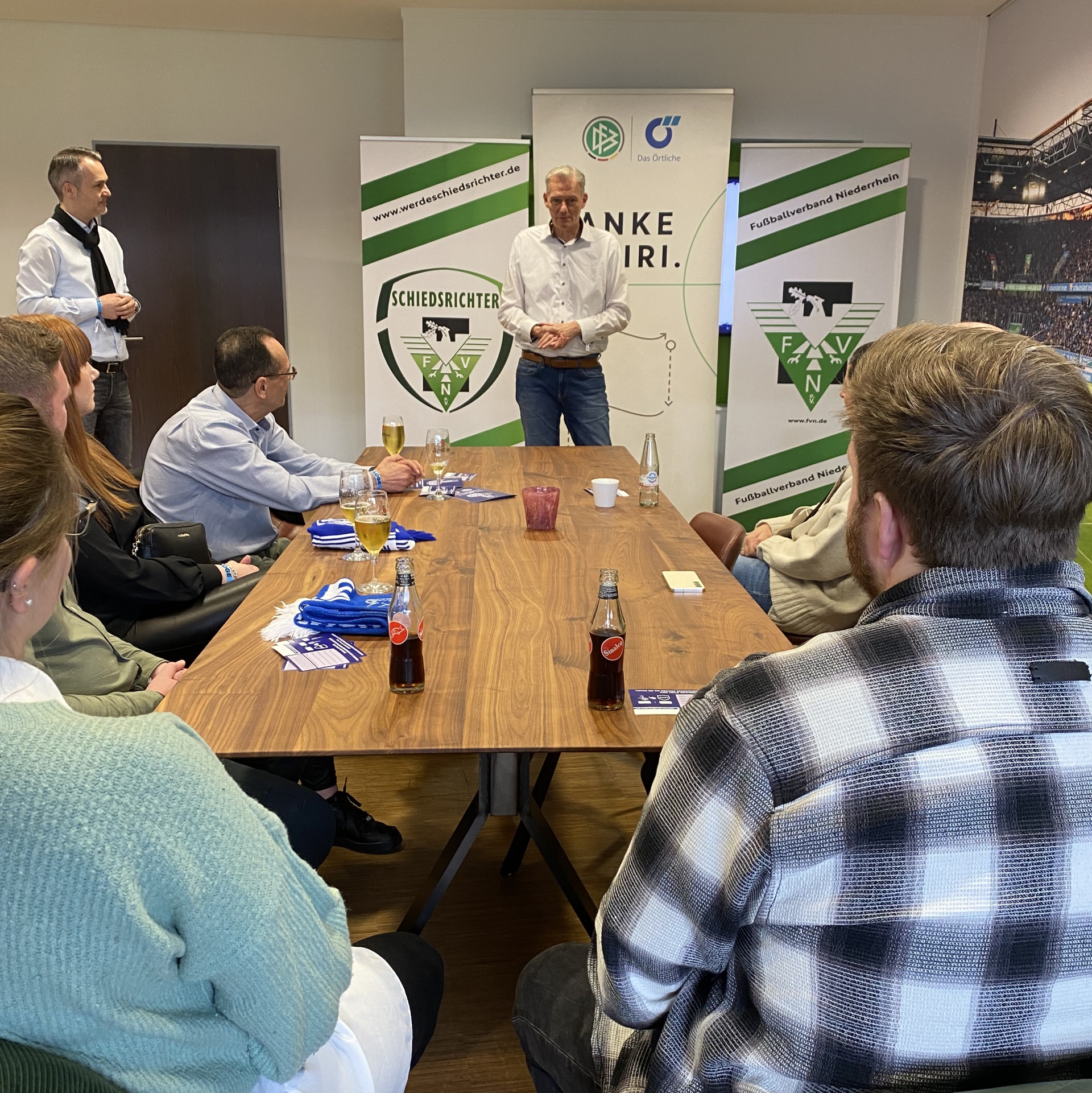 [{"clang_id":1,"value":"Gruppe diskutiert an einem großen Holztisch. Im Hintergrund Banner mit Fußballverein-Logo und Website. Personen sitzen und stehen, einige halten Getränke."}]