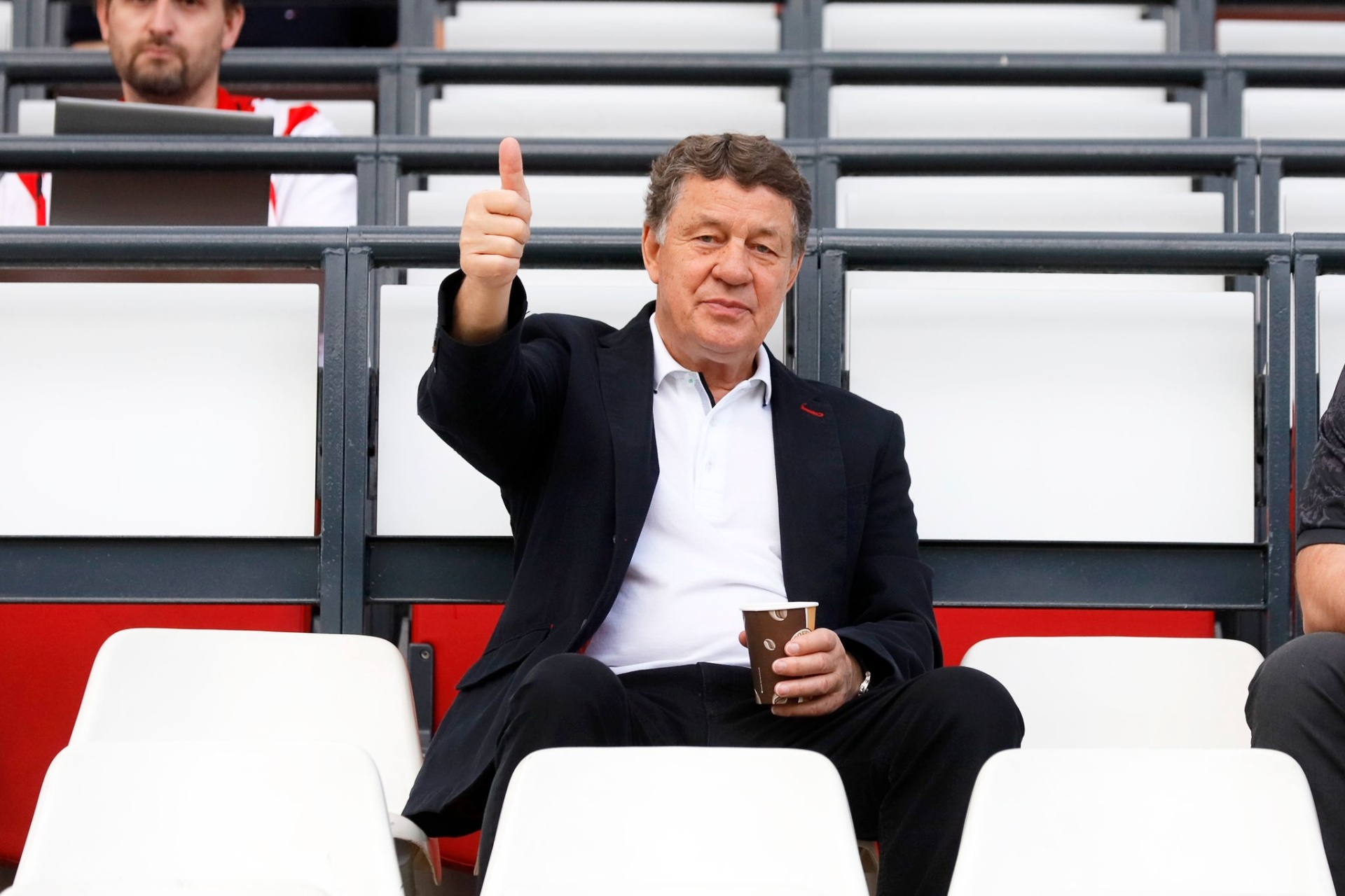 [{"clang_id":1,"value":"Mann in dunklem Anzug sitzt auf Stadiontribüne, zeigt Daumen hoch und hält Becher in der Hand. Im Hintergrund weitere Personen und Sitzreihen."}]