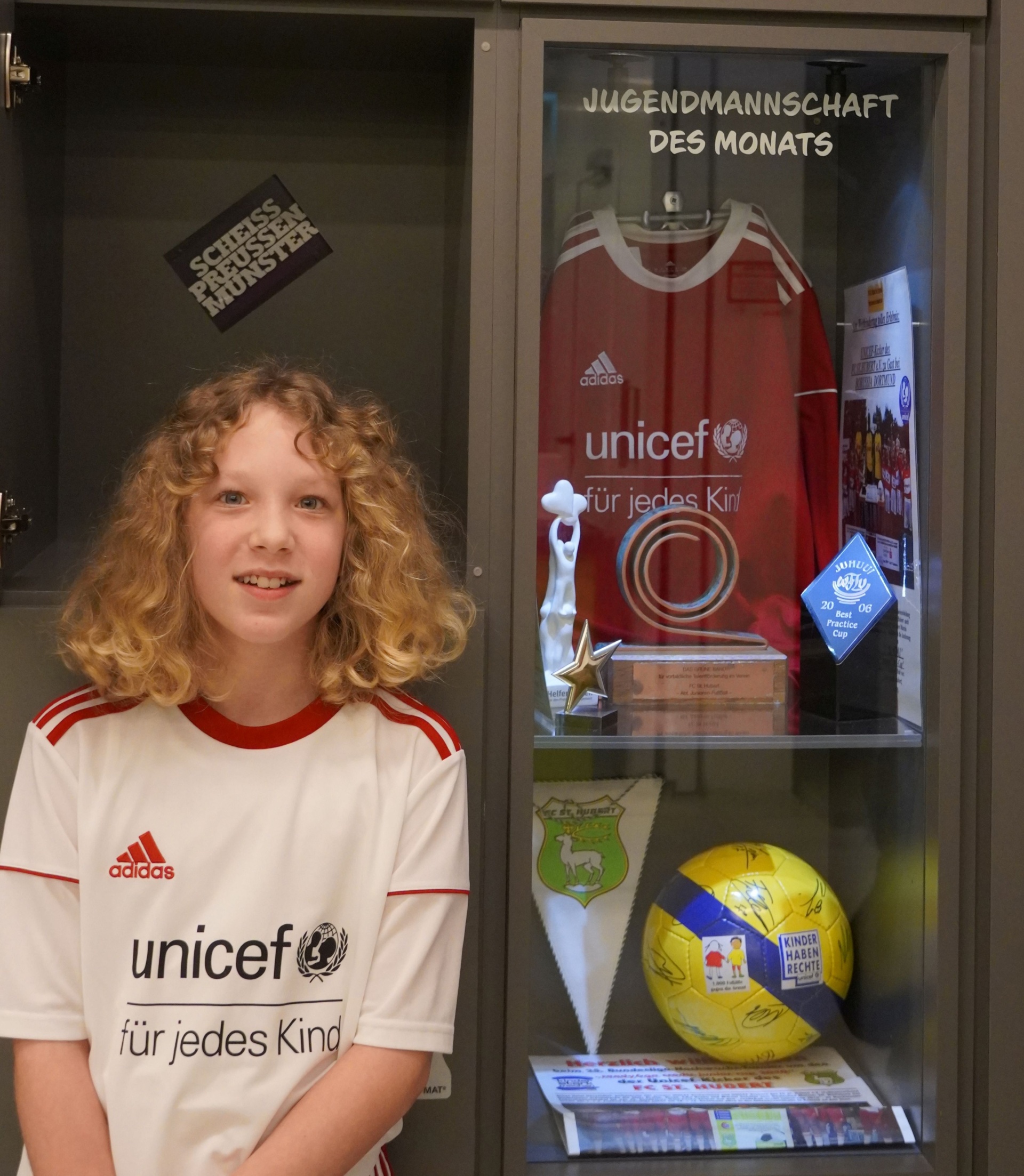 [{"clang_id":1,"value":"Junge mit lockigem Haar in weißem Fußballtrikot vor Vitrine. Vitrine zeigt Fußballausrüstung und Auszeichnungen, darunter Trikot mit UNICEF-Logo."}]