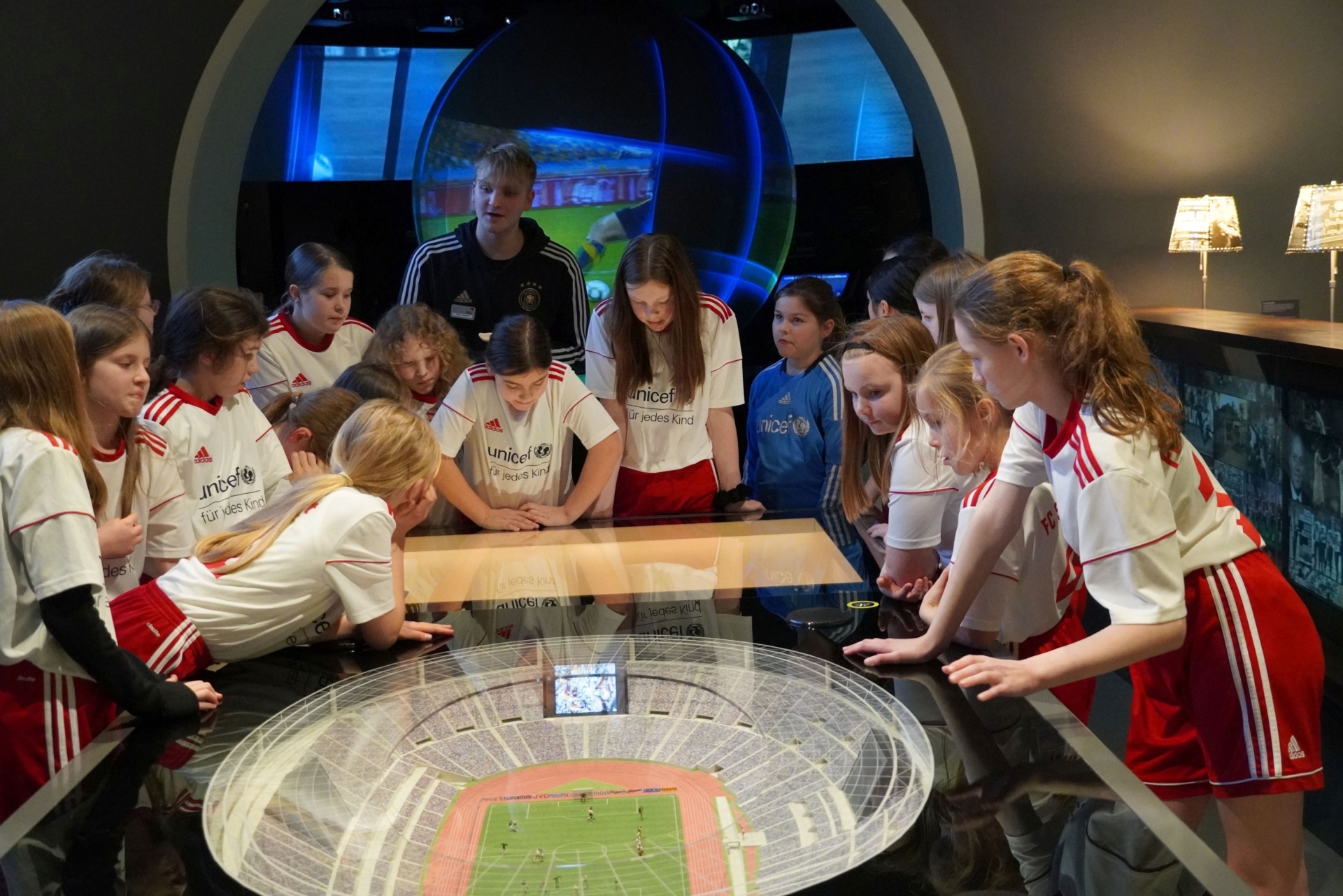 [{"clang_id":1,"value":"Gruppe Kinder in Fußballtrikots sitzt um einen runden Tisch mit integriertem Bildschirm, der ein Fußballfeld zeigt. Im Hintergrund ein Mann und Bildschirme."}]