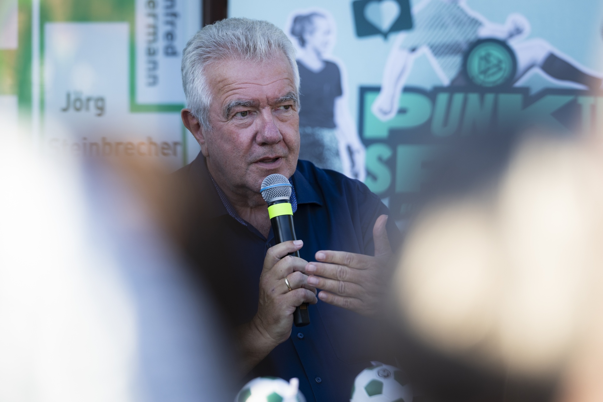 [{"clang_id":1,"value":"Mann mit Mikrofon spricht auf einer Veranstaltung. Im Hintergrund Banner mit Fußballmotiven und Namen."}]
