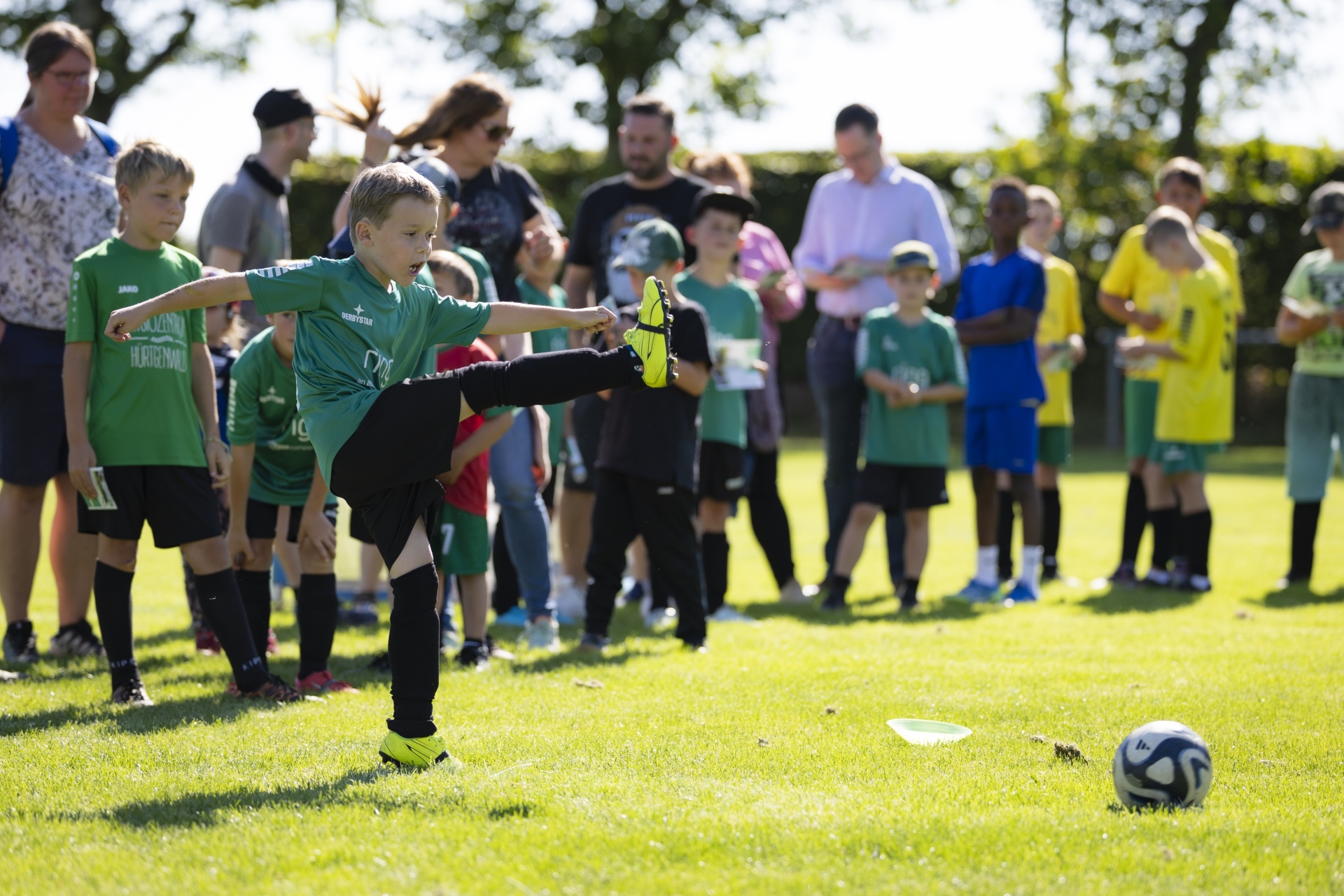 [{"clang_id":1,"value":"Junge im Fußballtrikot tritt auf einer Grasfläche einen Ball. Im Hintergrund stehen zuschauende Personen. Sonniger Tag."}]