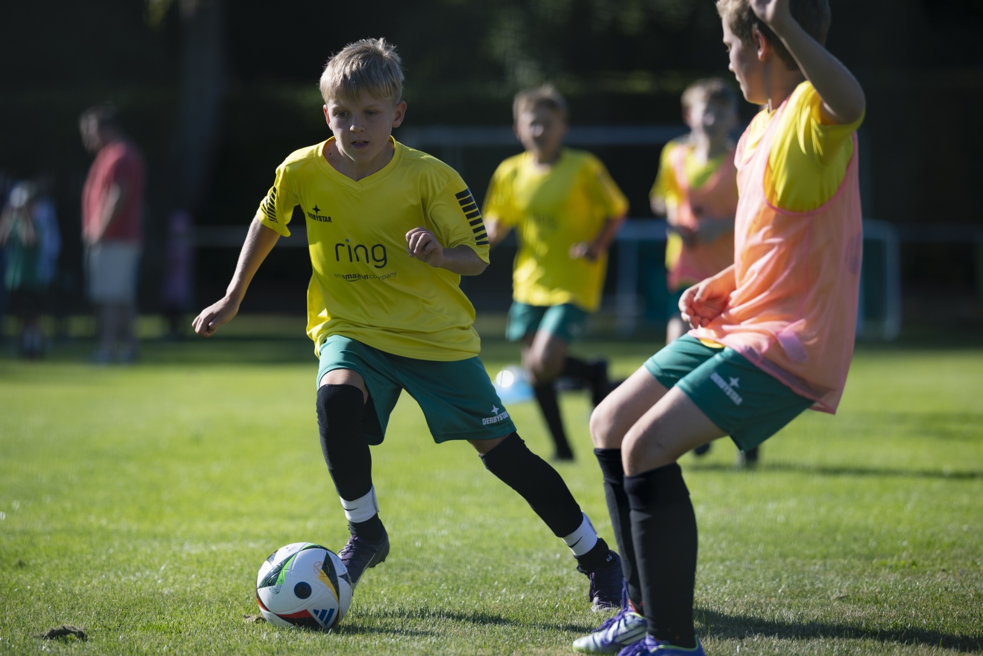 [{"clang_id":1,"value":"Fußballtraining: Junge mit Ball dribbelt an Gegenspieler. Im Hintergrund weitere Kinder beim Spielen auf Grünfläche."}]