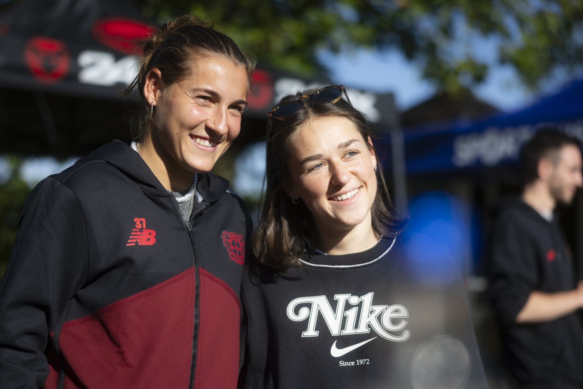 [{"clang_id":1,"value":"Zwei lächelnde Frauen posieren gemeinsam. Links trägt eine Frau eine New Balance Trainingsjacke, rechts eine Nike-Jacke. Im Hintergrund sind Menschen und Zelte zu sehen."}]
