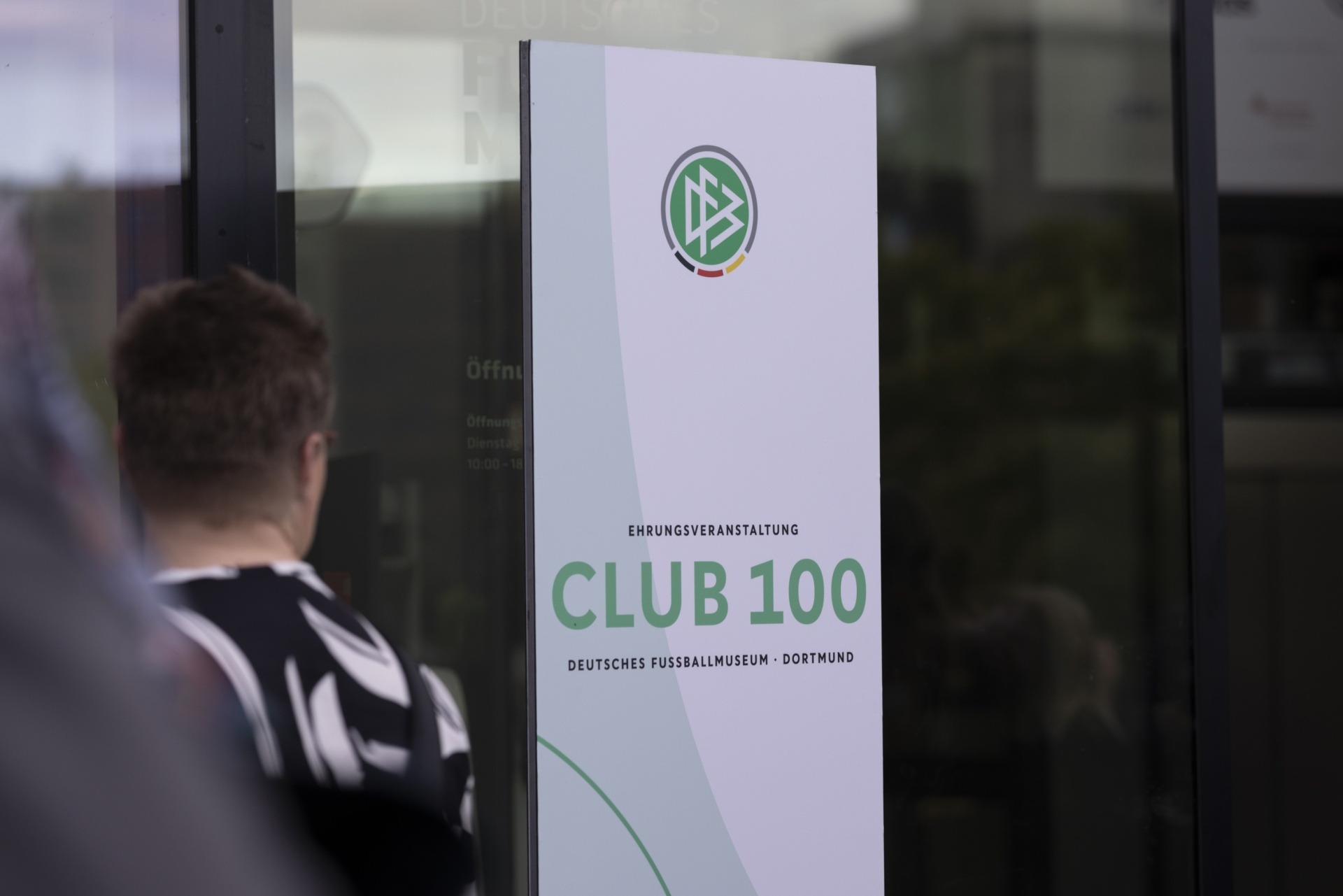 [{"clang_id":1,"value":"Fußballmuseum Dortmund: Plakat \"CLUB 100\" mit Vereinslogo. Links im Hintergrund ein Fußballspieler mit schwarz-weiß gestreiftem Trikot, durch Glas."}]