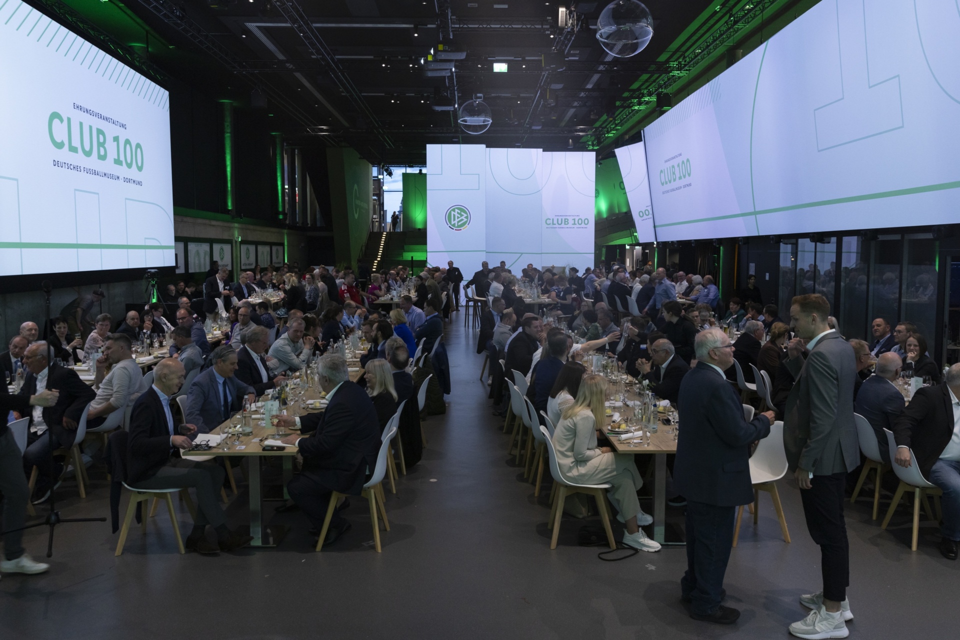 [{"clang_id":1,"value":"Großer Konferenzraum mit langen Tafeln, voll mit Menschen bei einem Dinner. Im Hintergrund eine Bühne mit dem Schriftzug \"CLUB 100\"."}]