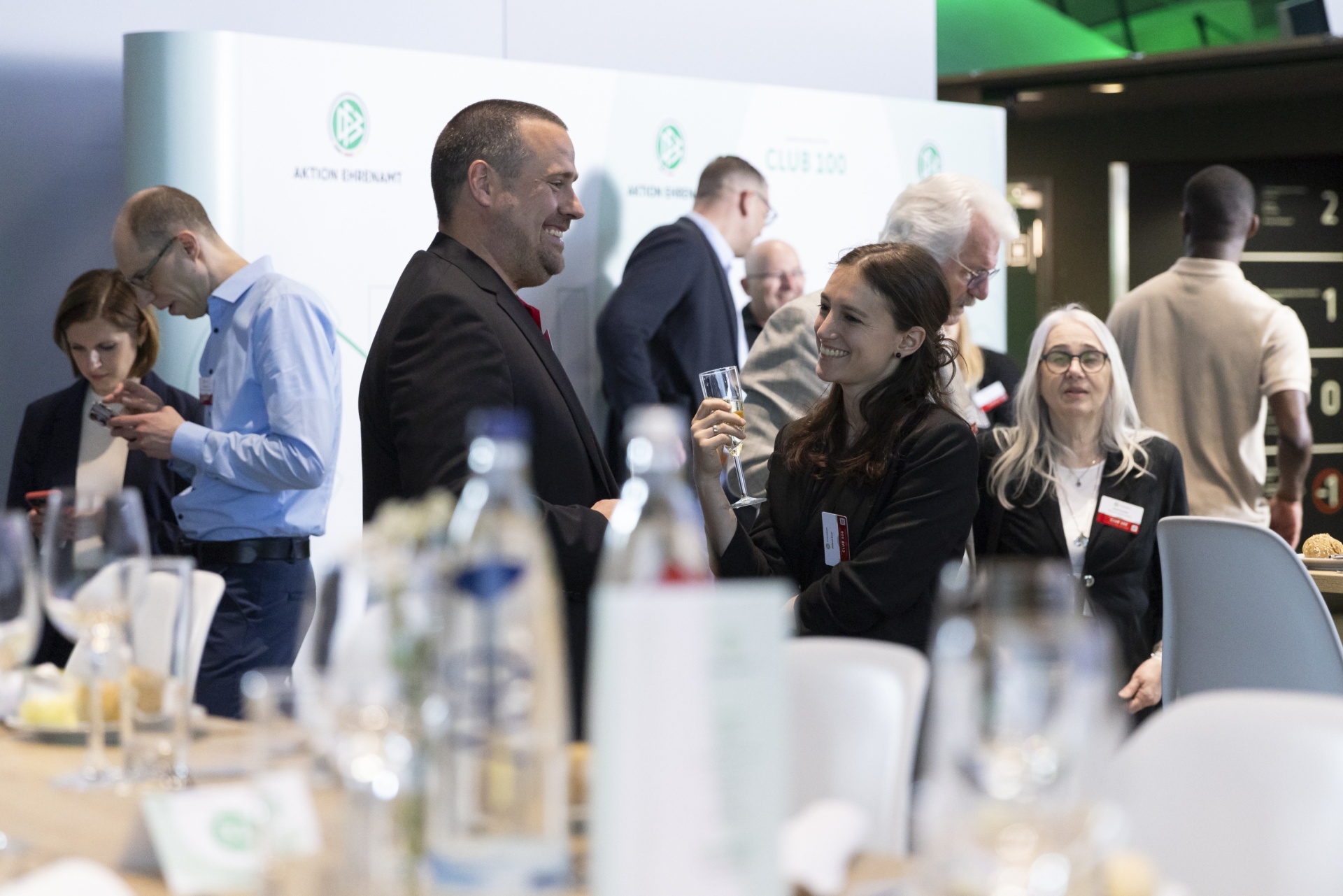 [{"clang_id":1,"value":"Gesellschaft in heller Halle. Menschen in Business-Kleidung unterhalten sich bei Getränken und Häppchen an Tischen. Im Hintergrund Banner mit Schriftzügen."}]