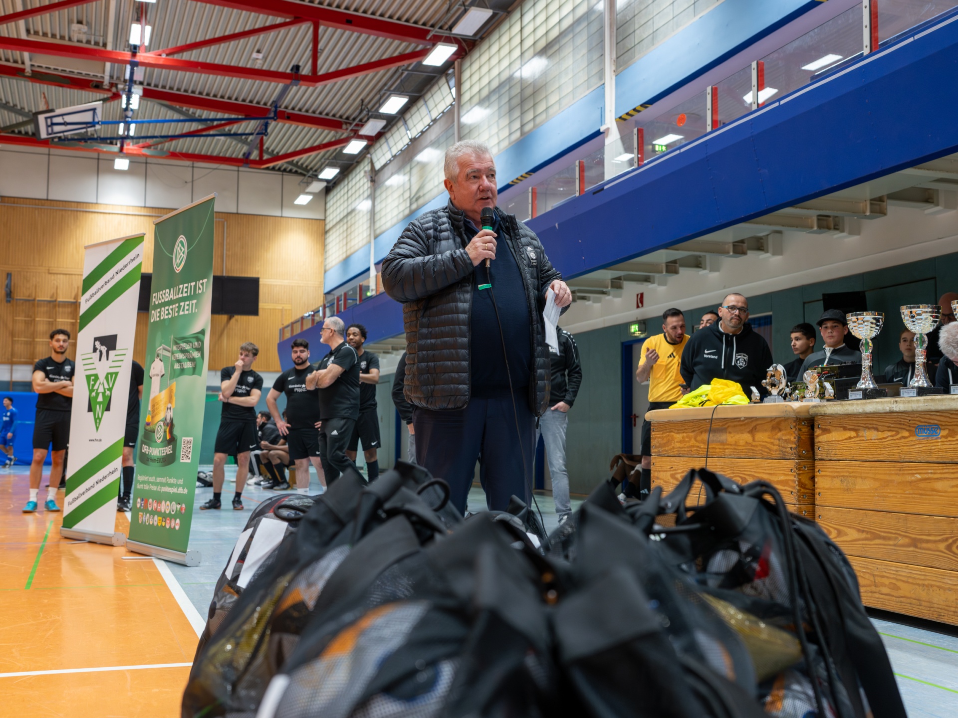 [{"clang_id":1,"value":"Halle mit Sportboden. Mann hält Rede vor Gruppe. Im Hintergrund Trophäensammlung, Banner und spielende Personen. Im Vordergrund Sporttaschen."}]