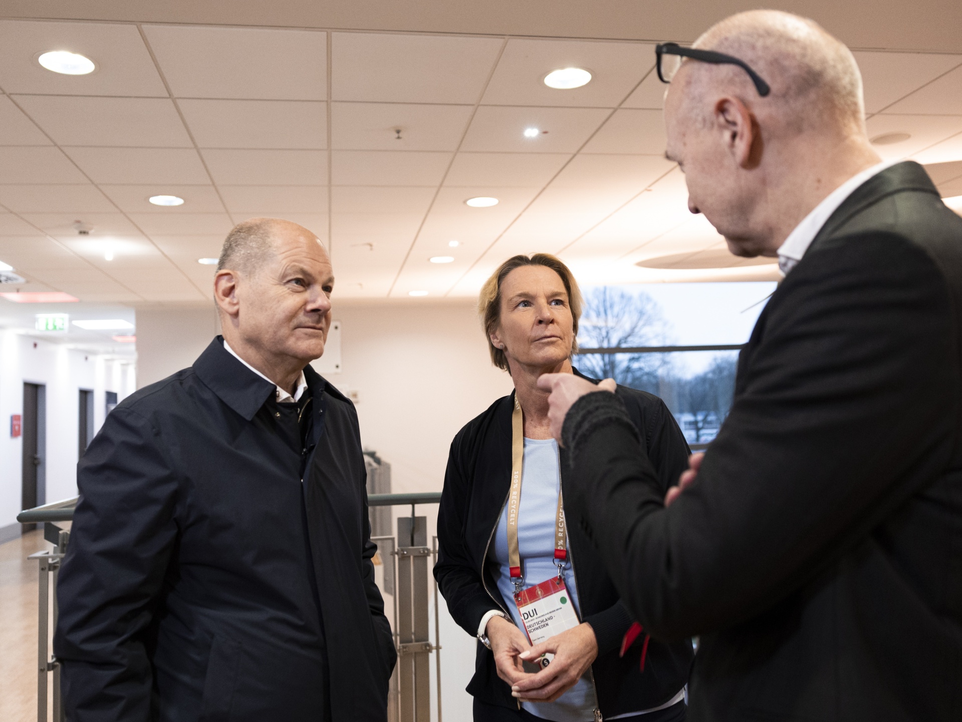 [{"clang_id":1,"value":"Drei Personen in einem hellen Innenraum im Gespräch. Links ein Mann mit dunklem Mantel, in der Mitte eine Frau mit Namensschild, rechts ein Mann im Anzug."}]