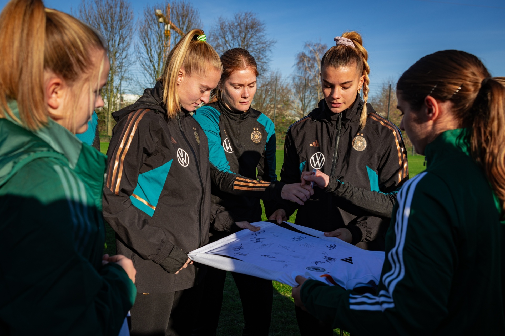 [{"clang_id":1,"value":"Fünf Fußballspielerinnen in Trainingsanzügen stehen auf einem Rasenfeld um einen Plan oder ein Dokument gebückt und besprechen etwas."}]