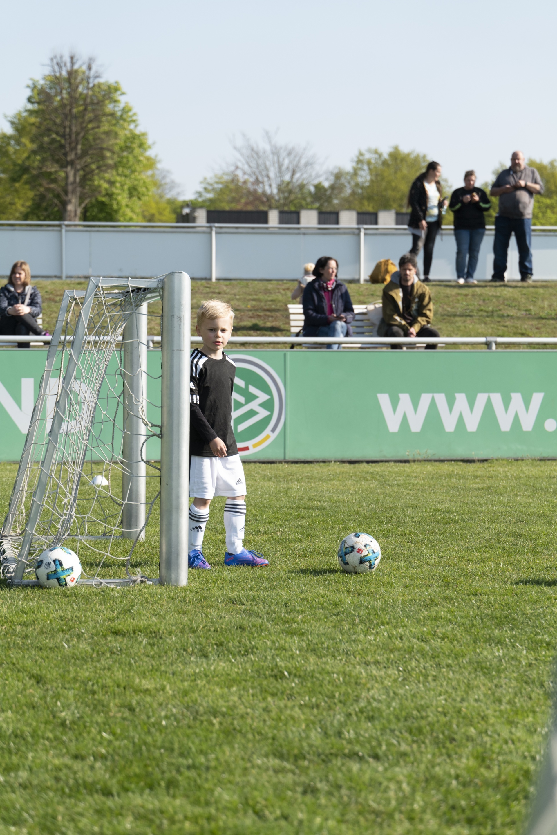 [{"clang_id":1,"value":"Junge steht auf grünem Fußballfeld vor kleinem Tor. Zwei Fußbälle liegen vor ihm. Im Hintergrund eine Bande mit Schriftzug und Zuschauer."}]