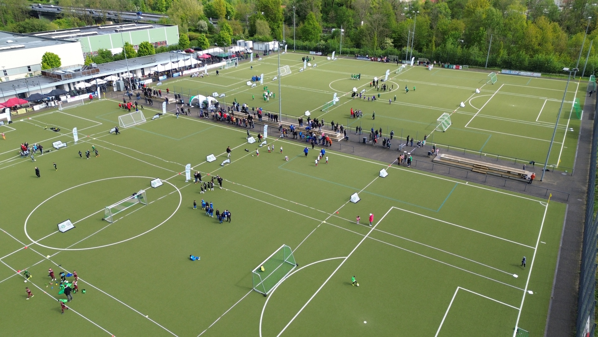 [{"clang_id":1,"value":"Luftaufnahme eines Fußballgeländes mit mehreren kleinen Spielfeldern. Spieler und Zuschauer sind zu sehen. Im Hintergrund Gebäude und Bäume."}]