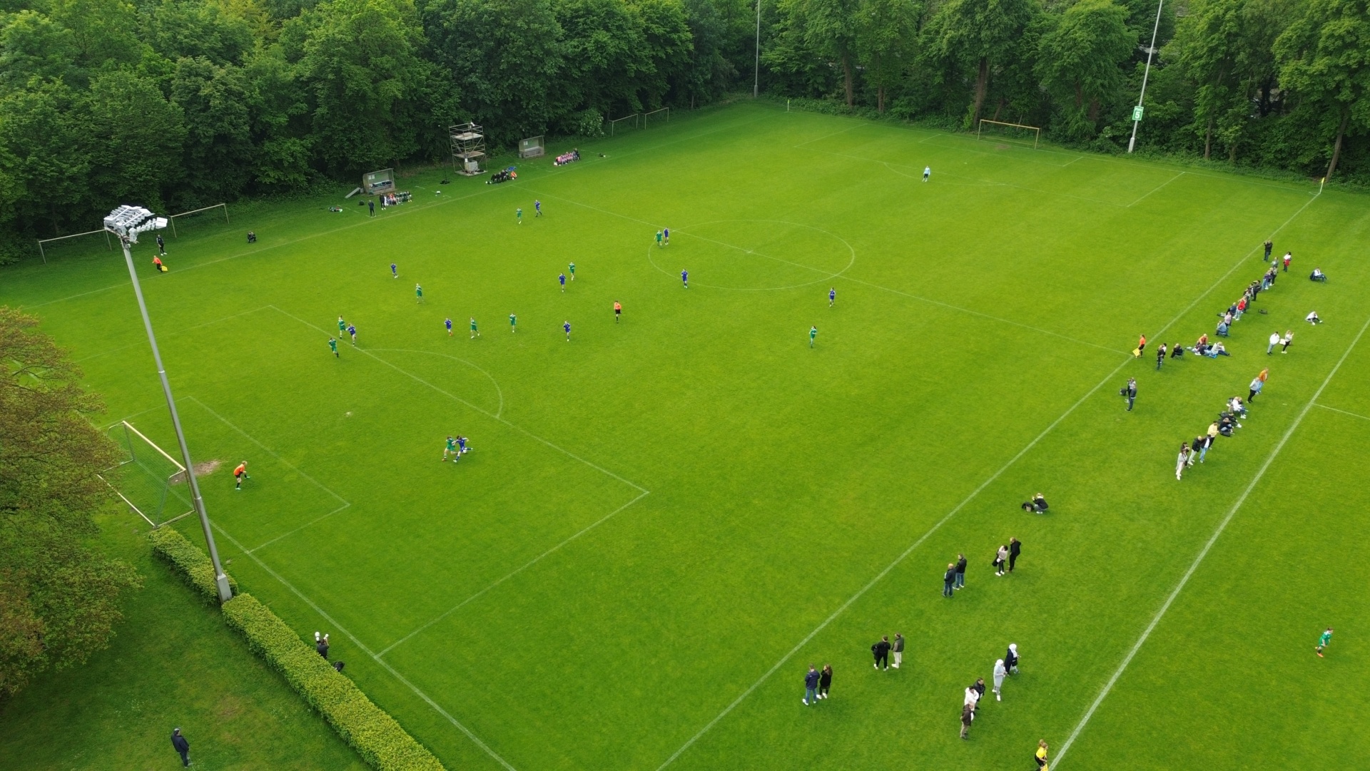 [{"clang_id":1,"value":"Fußballfeld mit spielenden Mannschaften und Zuschauern. Bäume säumen den Rand des Feldes, ein Flutlichtmast steht am Spielfeldrand."}]