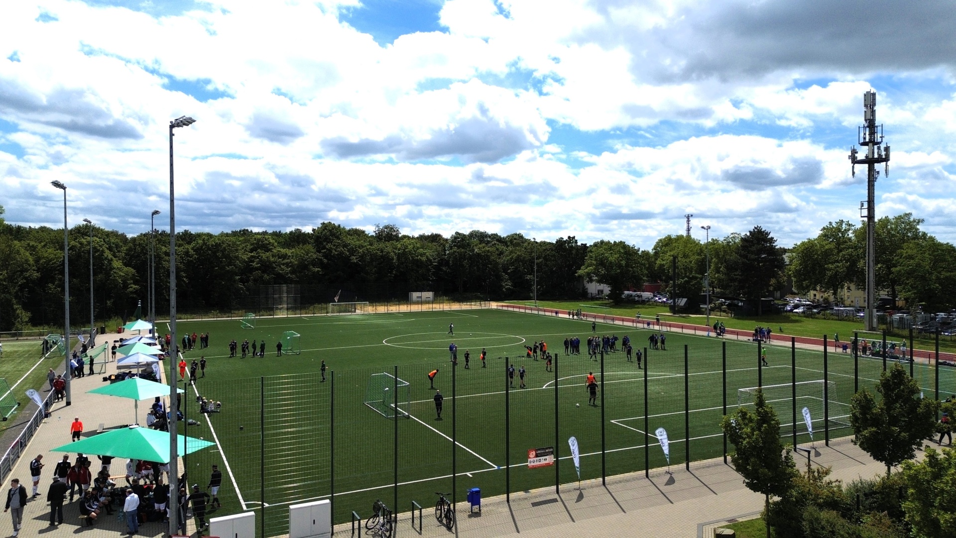 [{"clang_id":1,"value":"Fußballfeld mit zwei laufenden Spielen, umgeben von Zuschauern unter Sonnenschirmen und Bäumen. Im Hintergrund ein Mobilfunkmast."}]