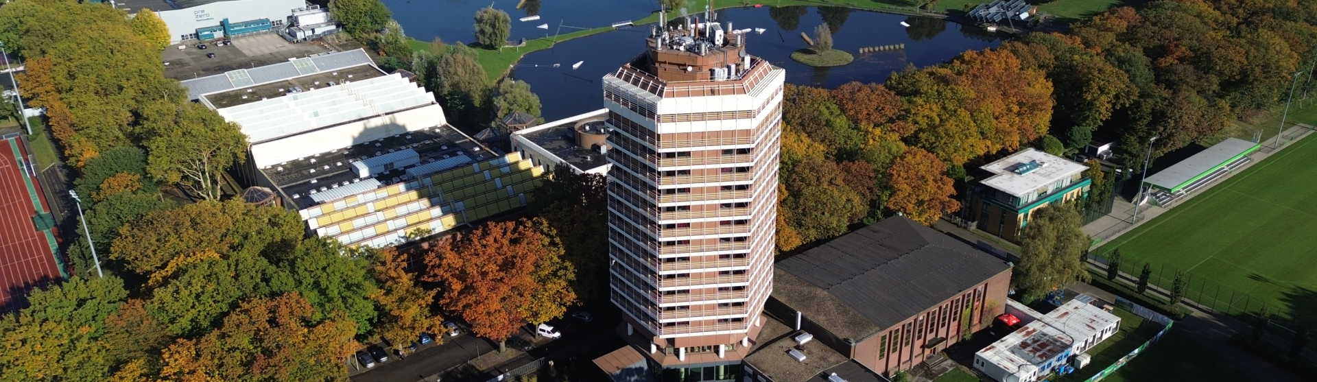 [{"clang_id":1,"value":"Hochhaus mit bräunlicher Fassade inmitten von Gebäuden, Bäumen und einer Wasserfläche. Sportplatz im Hintergrund rechts."}]