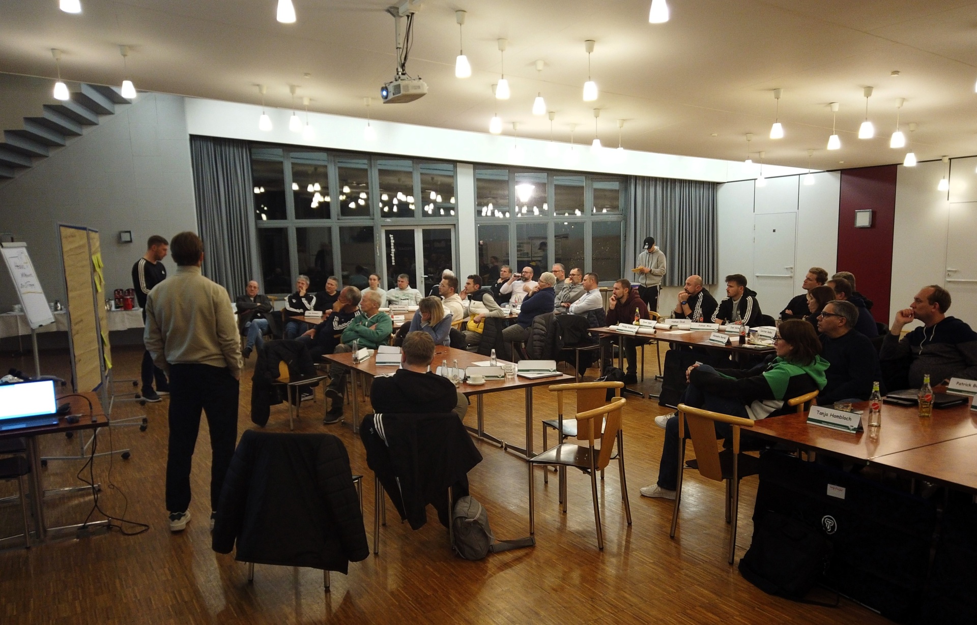 [{"clang_id":1,"value":"Großer Raum mit Konferenztisch und etwa 30 teilnehmenden Personen bei einer Besprechung. Im Hintergrund Fenster mit Außenansicht und Lichtquellen an der Decke."}]