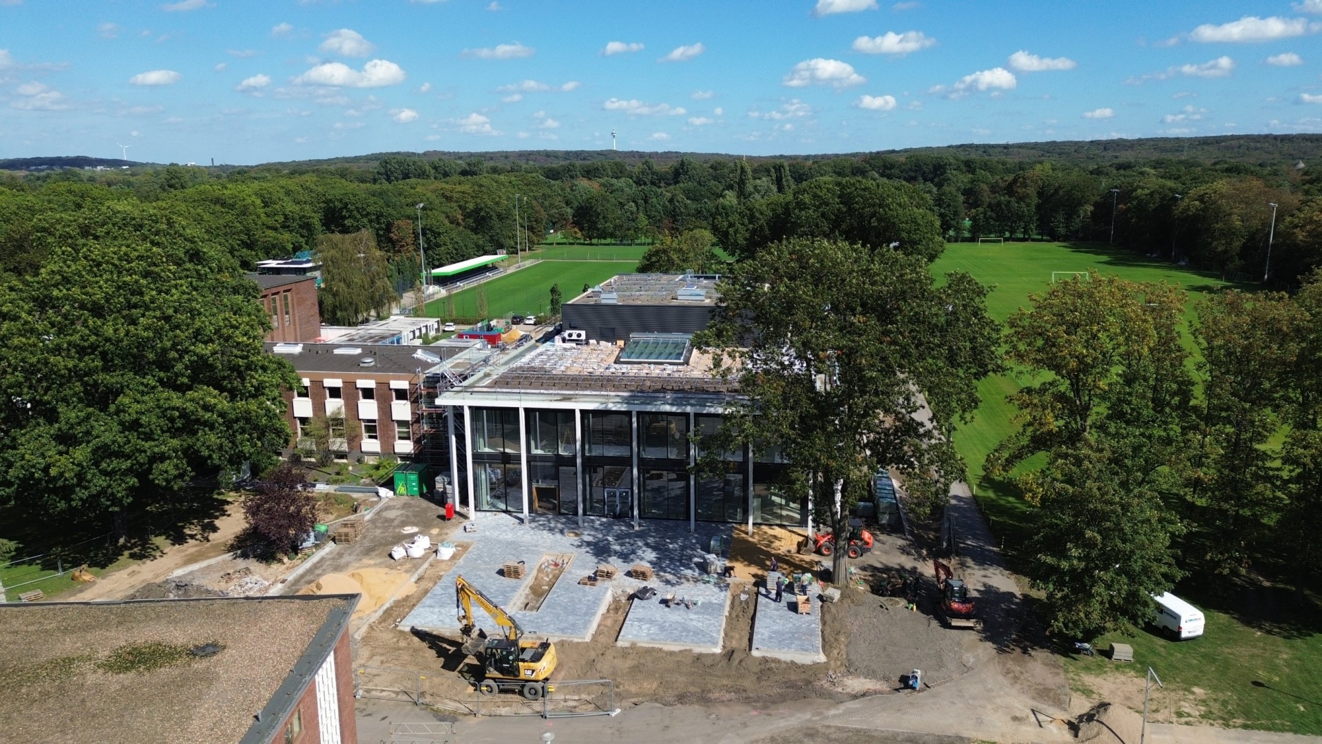 Luftaufnahme einer Baustelle neben einem mehrstöckigen Gebäude. Im Hintergrund ein Sportplatz mit Rasenfeld und umgebende Bäume unter blauem Himmel.