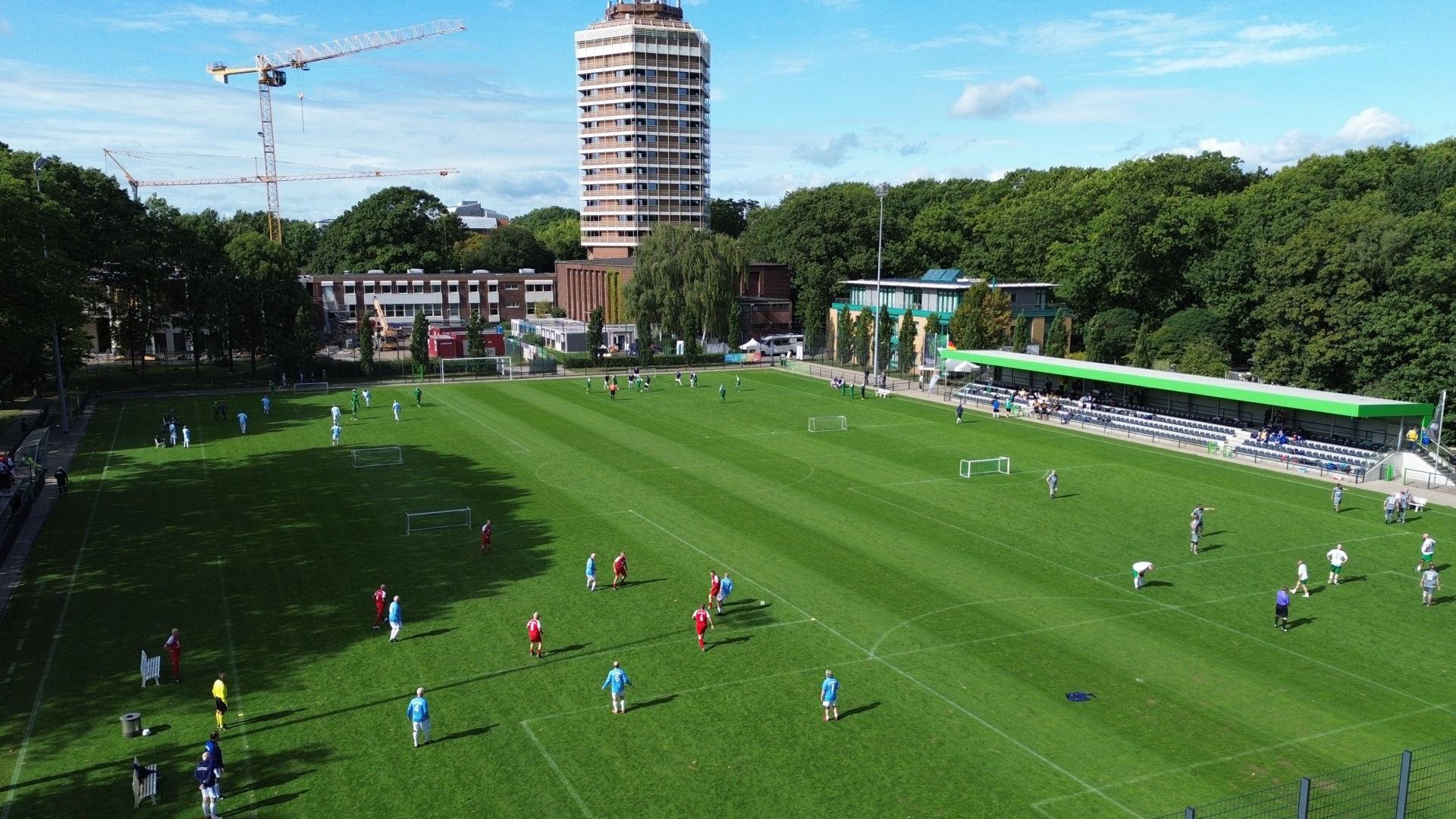 [{"clang_id":1,"value":"Fußballfeld mit Spielern in blau-weißen und roten Trikots. Im Hintergrund Gebäude, Bäume und ein Bau Kran."}]