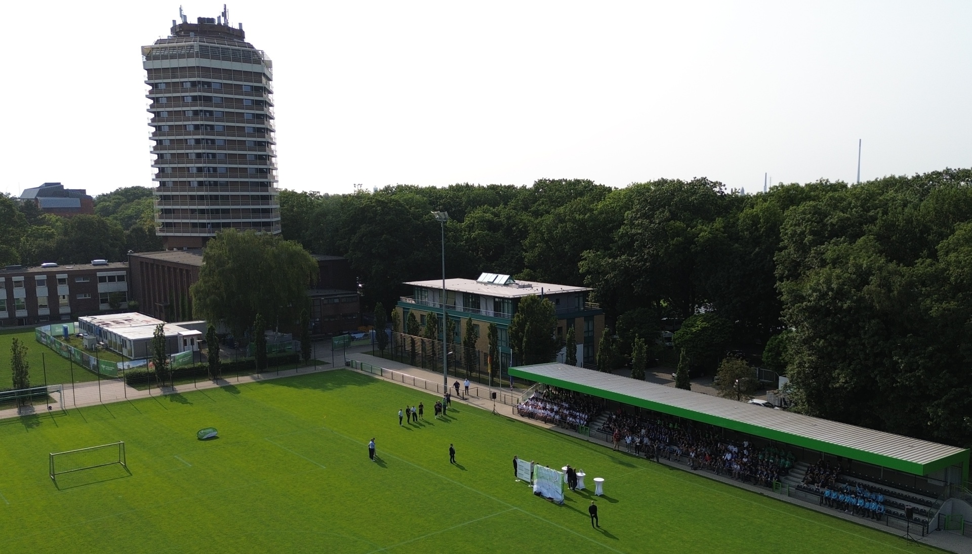 [{"clang_id":1,"value":"Fußballfeld mit spielenden Personen, dahinter ein niedriges Gebäude mit Tribüne und ein hoher Wohnturm im Hintergrund. Bäume säumen den Rand des Geländes."}]
