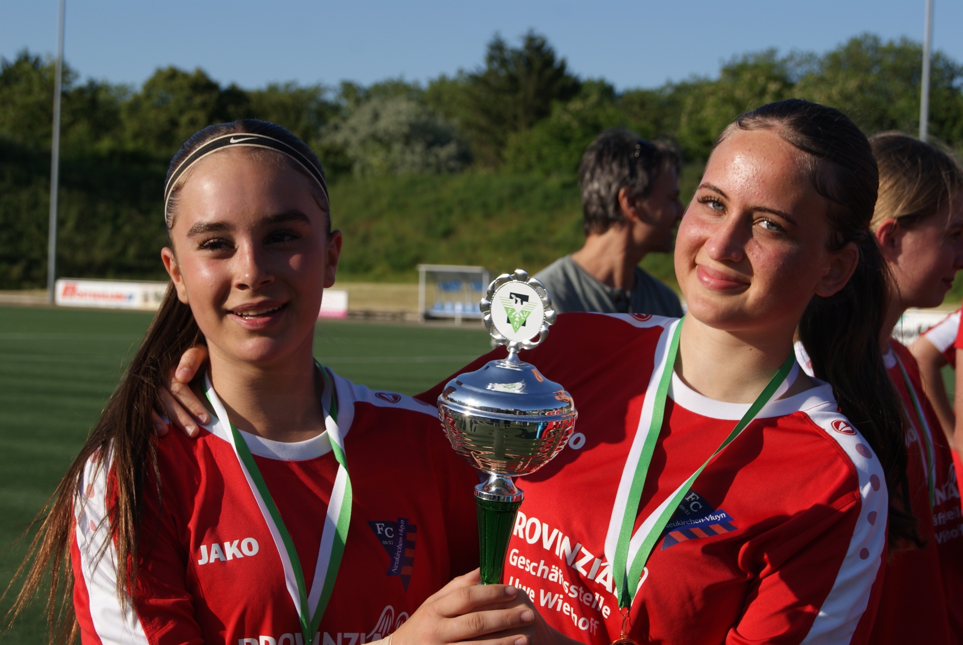 [{"clang_id":1,"value":"Zwei junge Fußballspielerinnen mit Medaillen und Pokal stehen lächelnd vor einem Fußballfeld. Beide tragen rote Trikots mit weißen Details."}]