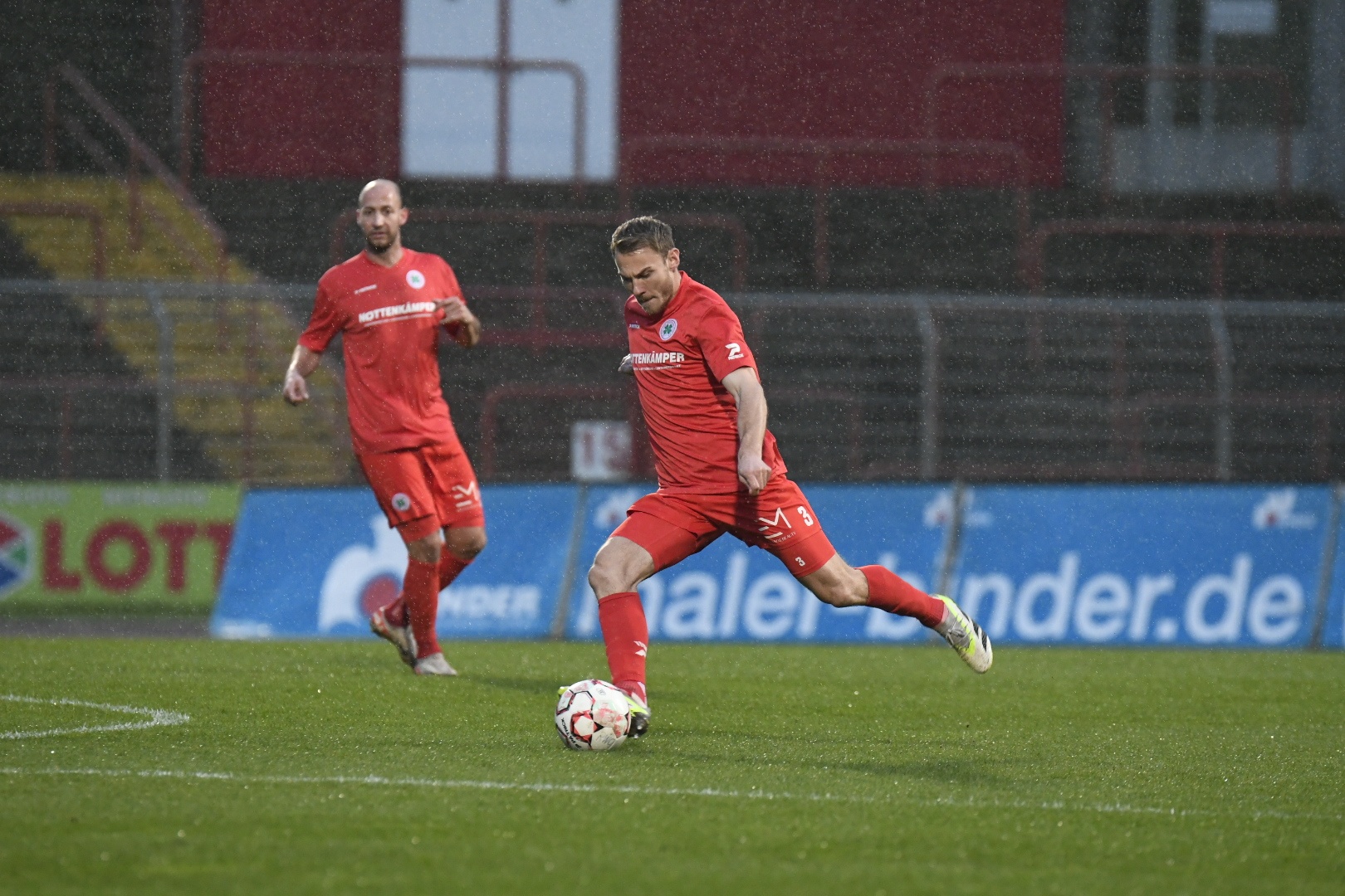 [{"clang_id":1,"value":"Zwei Fußballspieler in roten Trikots auf grünem Rasen bei Regen. Einer schießt den Ball, der andere steht daneben. Im Hintergrund eine Tribüne."}]