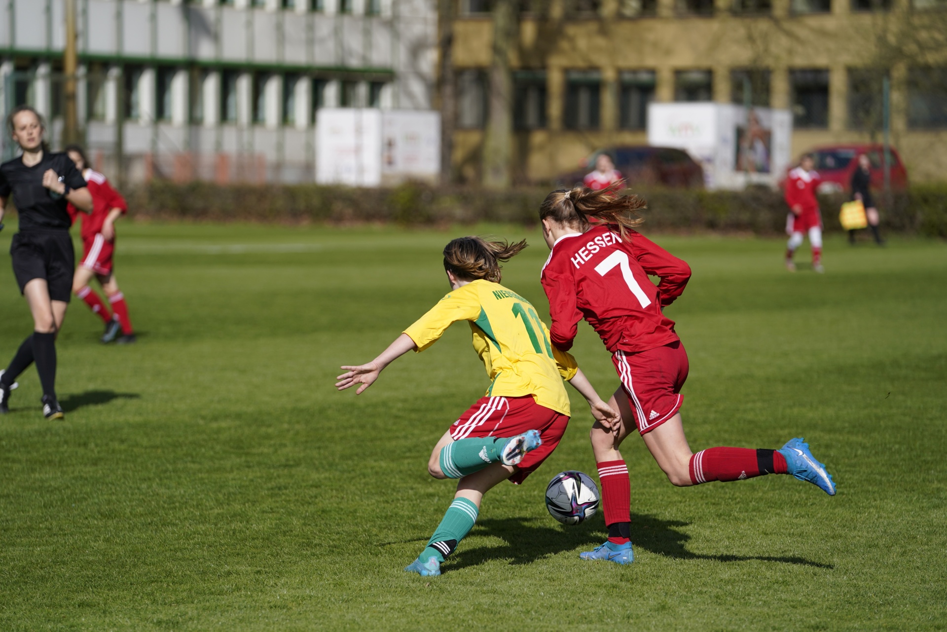 [{"clang_id":1,"value":"Zwei Fußballspielerinnen im Spiel. Eine trägt rot mit Nummer 7, die andere gelb-grün. Im Hintergrund Gebäude und weitere Spieler. Rasenplatz."}]
