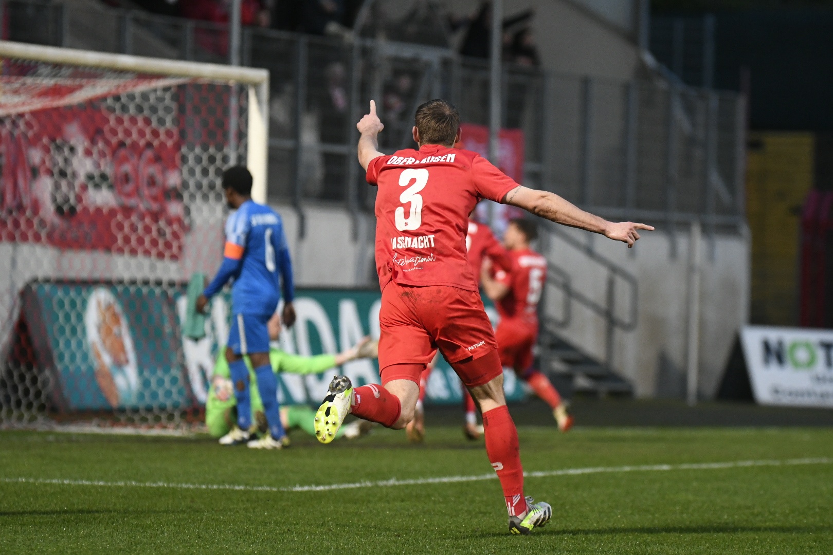 [{"clang_id":1,"value":"Fußballspieler in rotem Trikot mit Nummer 3 jubelt, Arme hoch, Finger zeigt nach oben. Im Hintergrund weitere Spieler und ein Fußballtor."}]
