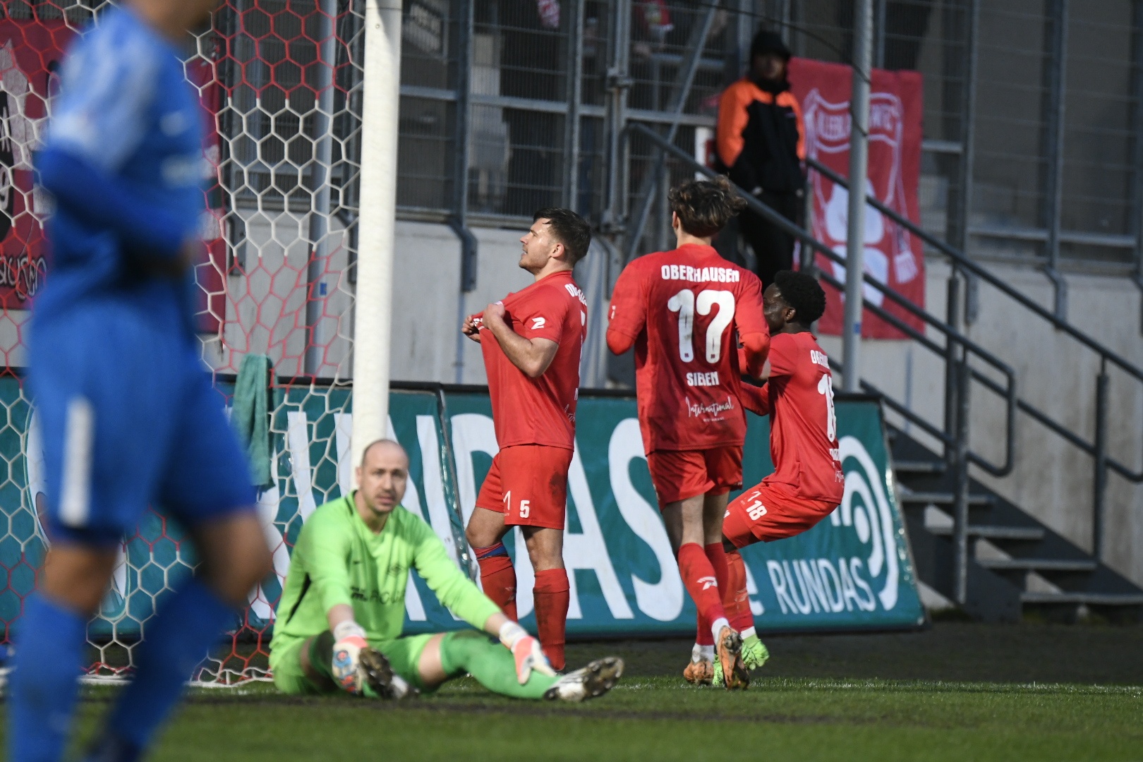 [{"clang_id":1,"value":"Fußballspiel: Roter Spieler jubelt mit Teamkollegen vor dem Tor. Im Hintergrund steht ein blauer Spieler und der Torwart kniet am Boden."}]