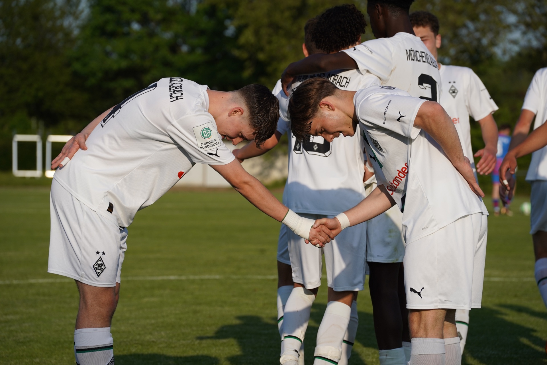 [{"clang_id":1,"value":"Fußballspieler in weißen Trikots und grünen Shorts stehen auf einem Rasenfeld. Zwei Spieler verneigen sich gegenseitig und fassen sich an den Unterarmen. Im Hintergrund weitere Spieler."}]