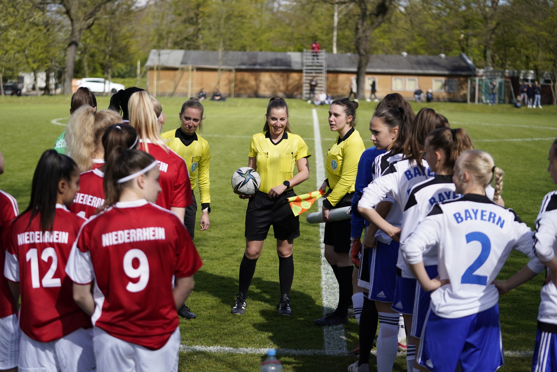 [{"clang_id":1,"value":"Fußballspiel: Schiedsrichterin mit zwei Mannschaften im Kreis, rote und weiße Trikots, auf grünem Rasen. Im Hintergrund Gebäude und Bäume."}]