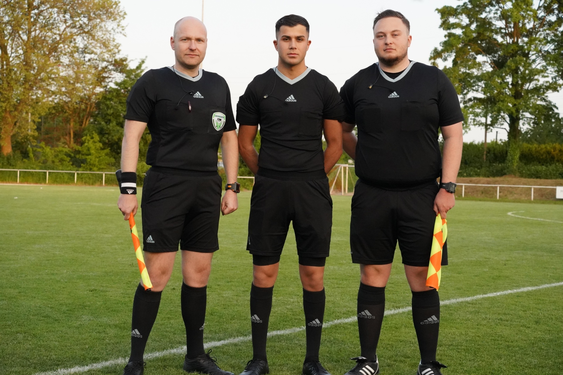 [{"clang_id":1,"value":"Drei Fußballschiedsrichter in schwarzer Sportbekleidung stehen auf einem Fußballfeld. Links und rechts halten sie gelb-schwarze Fahnen."}]
