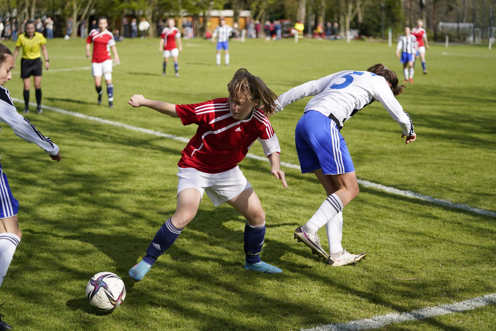 [{"clang_id":1,"value":"Fußballspiel: Jugendlicher in rotem Trikot dribbelt den Ball, wird von Gegenspieler in blauem Trikot bearbeitet. Hintergrund: weitere Spieler und Zuschauer."}]