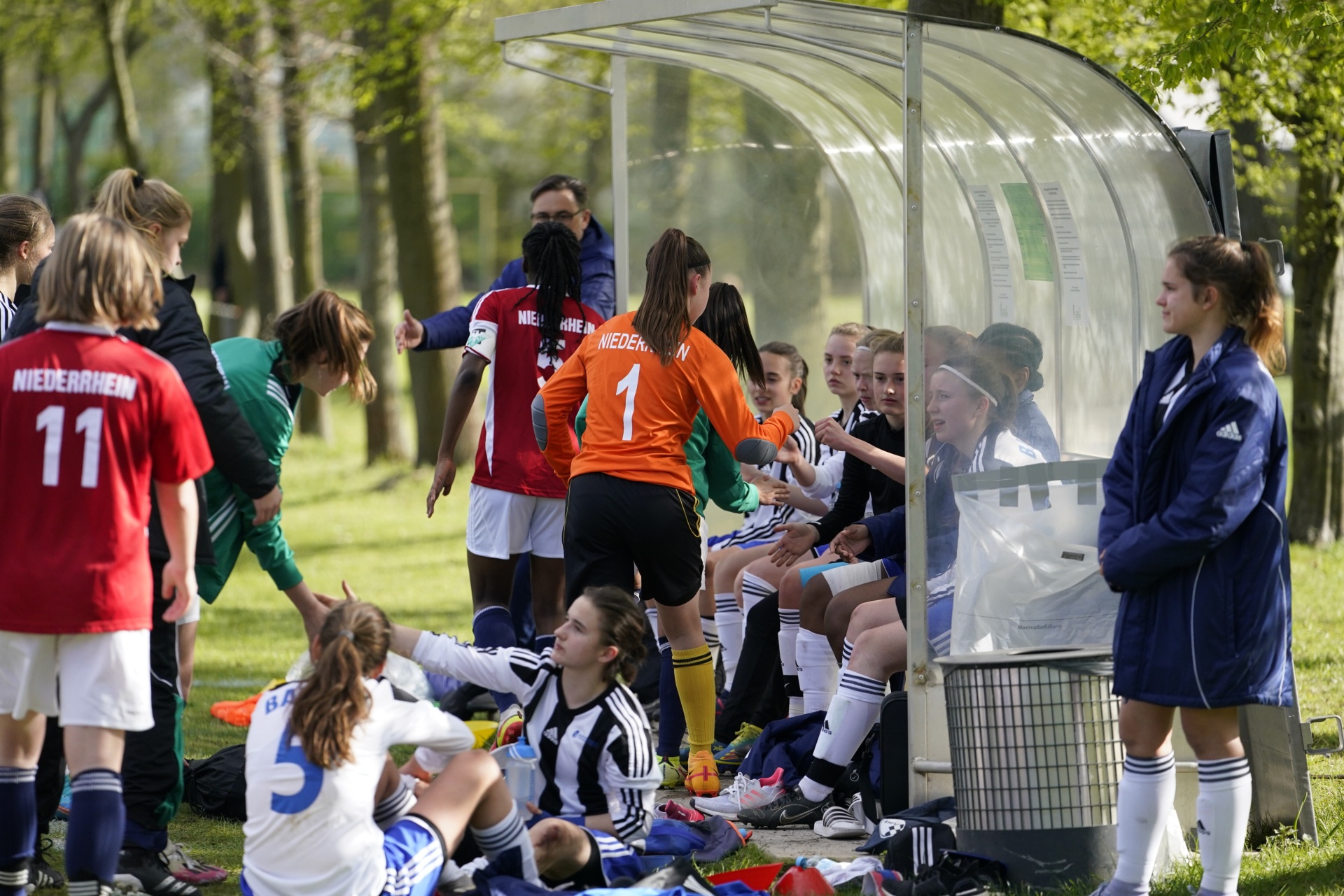 [{"clang_id":1,"value":"Fußballteam aus jungen Frauen/Mädchen auf einem Spielfeld. Einige sitzen/liegen am Spielfeldrand, andere stehen um eine Trainerin herum. Im Hintergrund Bäume und ein Spielerstand."}]