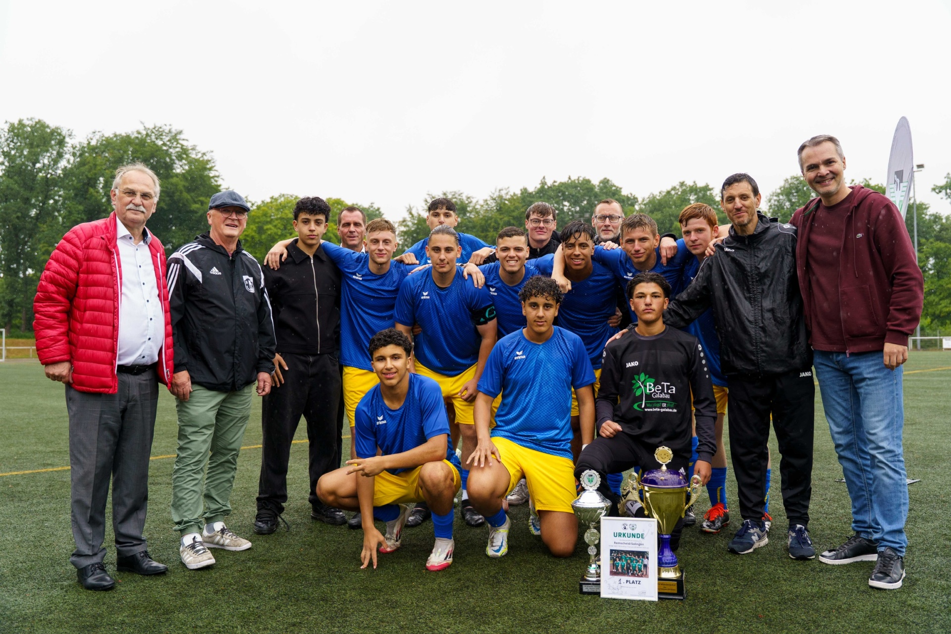 [{"clang_id":1,"value":"Fußballmannschaft in blau-gelben Trikots mit Trainern und Funktionären auf grünem Rasen, präsentieren Pokale und Urkunden."}]