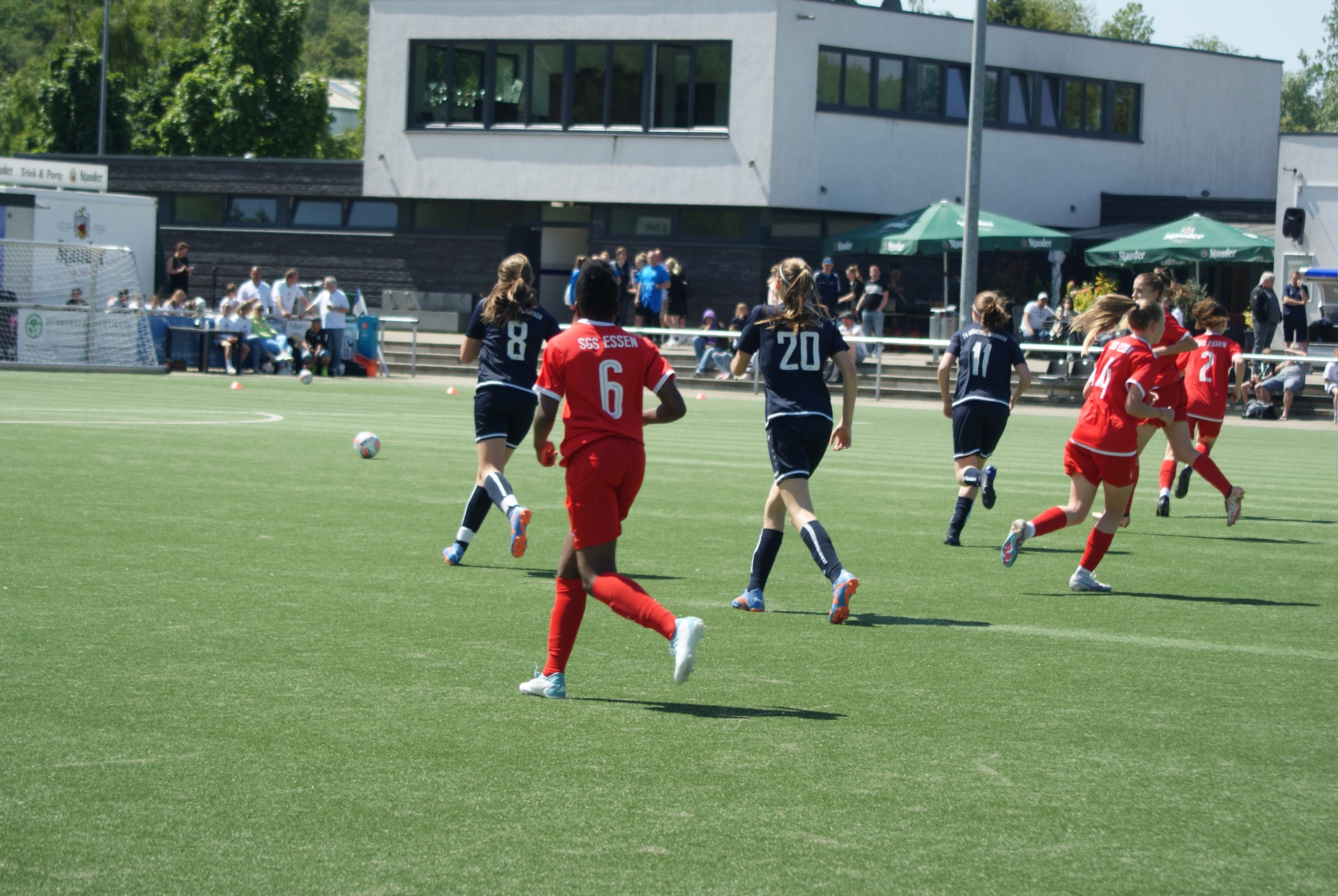 [{"clang_id":1,"value":"Fußballspiel auf grünem Rasen. Spielerinnen in roten und dunklen Trikots kämpfen um den Ball. Im Hintergrund Gebäude und Zuschauer."}]