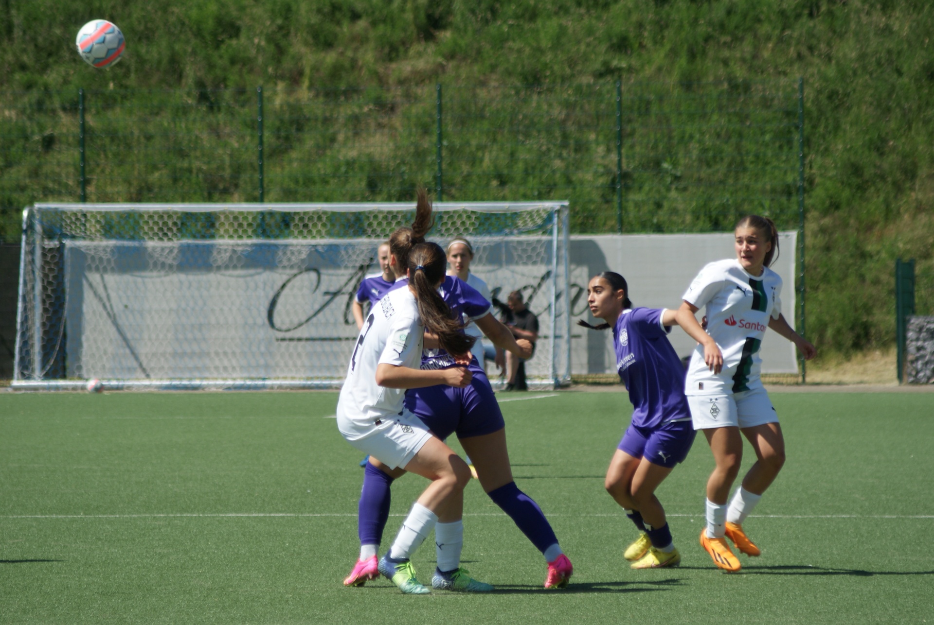 [{"clang_id":1,"value":"Fußballspiel mit vier jungen Frauen auf Kunstrasenplatz. Zwei Spielerinnen in Lila und zwei in Weiß/Grün kämpfen um den Ball. Im Hintergrund ein Fußballtor und Grünland."}]