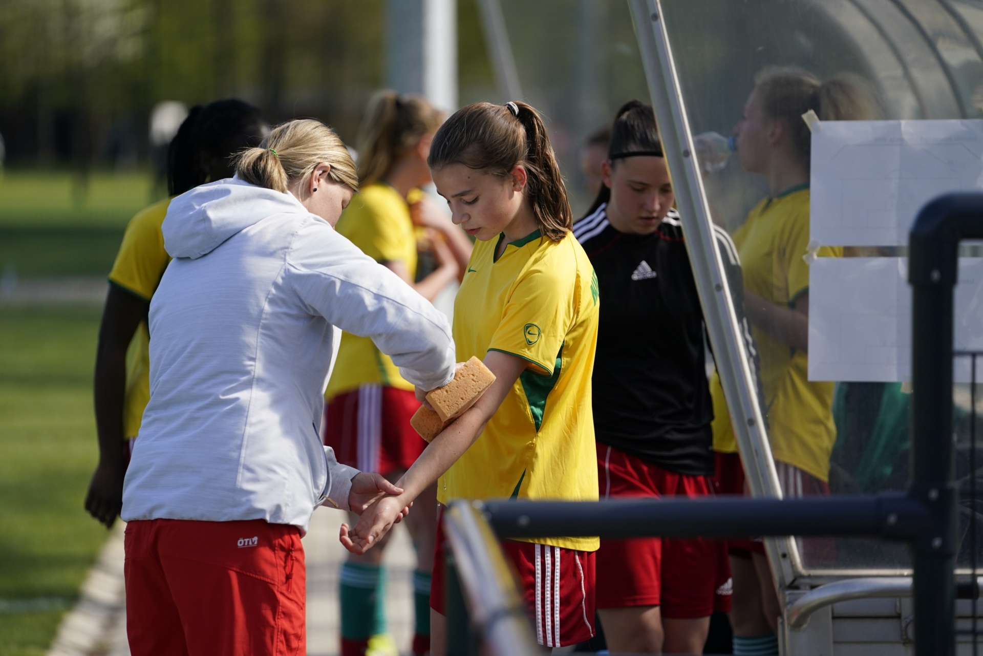 [{"clang_id":1,"value":"Trainerin versorgt Fußballspielerin am Bein. Im Hintergrund weitere Spielerinnen und ein Unterstand. Sportfeld im Hintergrund."}]