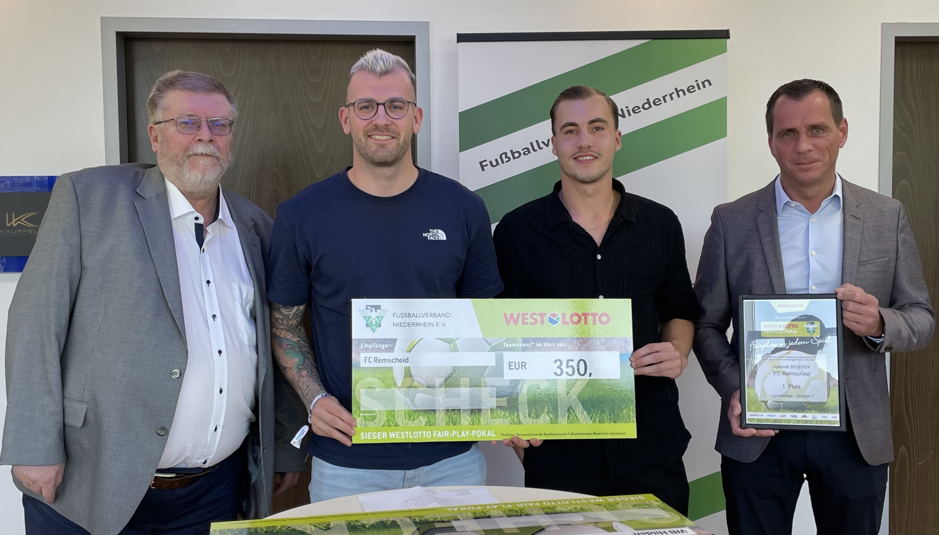 [{"clang_id":1,"value":"Vier Männer stehen nebeneinander und halten eine Scheck und eine Urkunde. Im Hintergrund ein Banner mit dem Schriftzug \"Fußballverband Niederrhein\"."}]