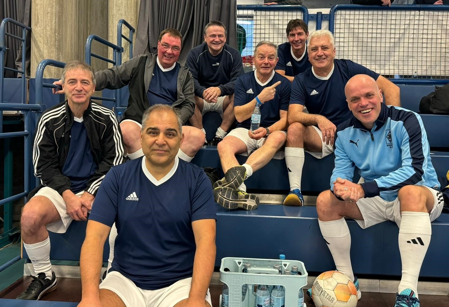 [{"clang_id":1,"value":"Fußballmannschaft sitzt auf einer Bank in einer Sporthalle. Sie tragen blaue und hellblaue Sportbekleidung. Vor ihnen stehen Wasserflaschen und ein Fußball."}]