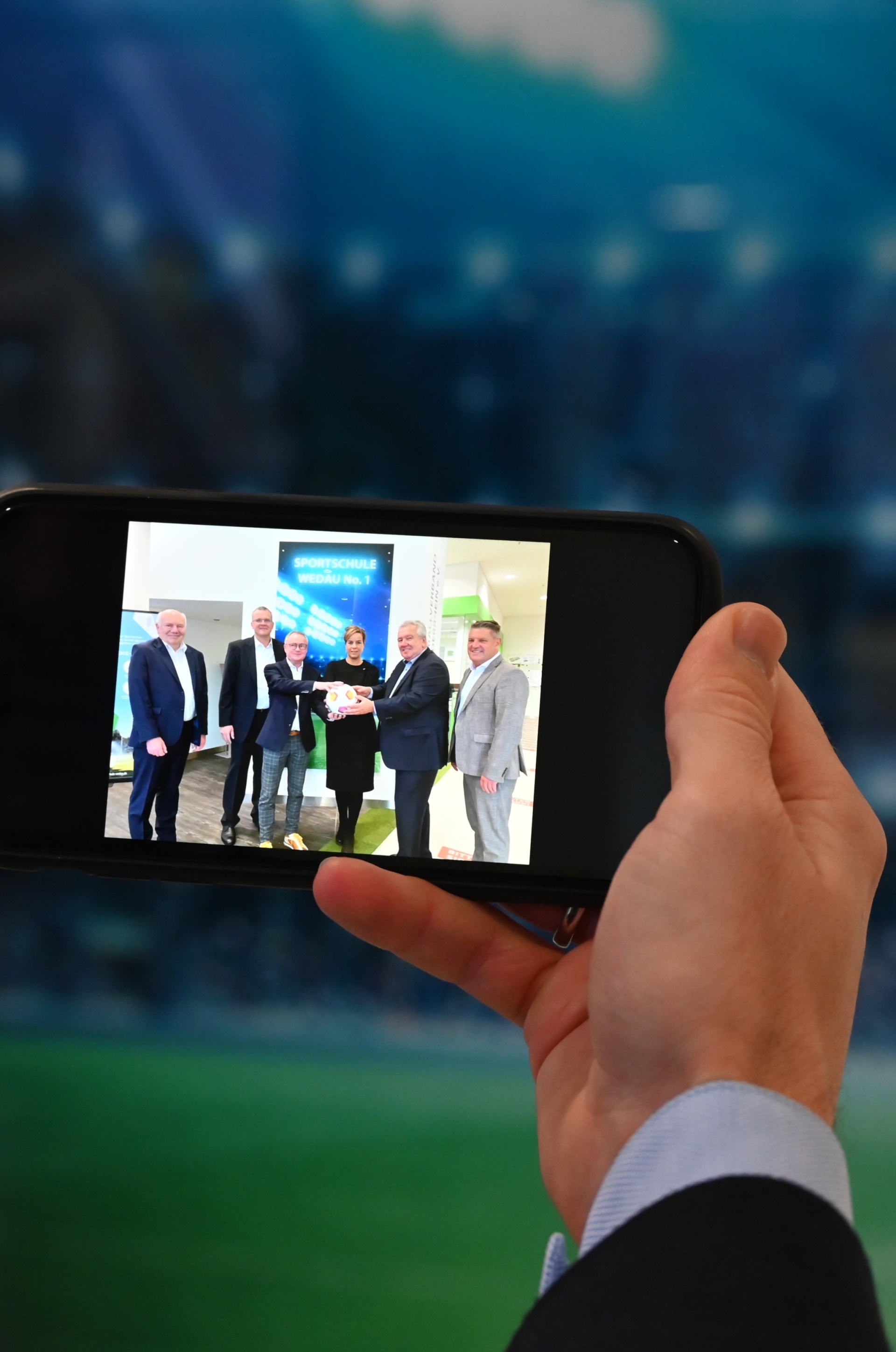[{"clang_id":1,"value":"Hand hält Smartphone, das ein Foto von vier Männern in Anzügen zeigt, die sich die Hände reichen. Hintergrund: Stadion."}]