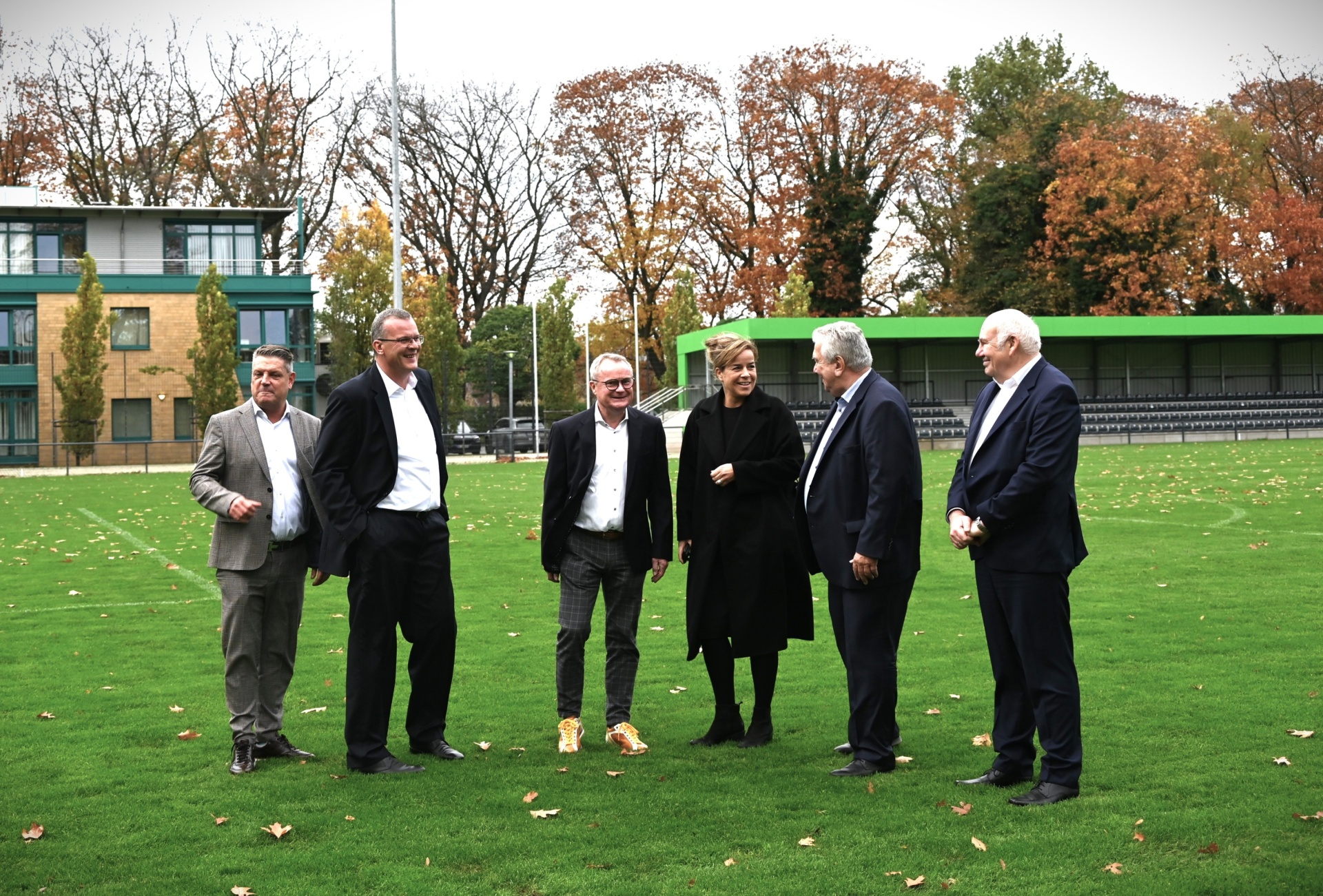 [{"clang_id":1,"value":"Sechs Männer und eine Frau stehen auf einem Rasenplatz vor einem Gebäude mit Zuschauertribünen und Bäumen im Herbstlaub. Alle tragen formelle Kleidung."}]
