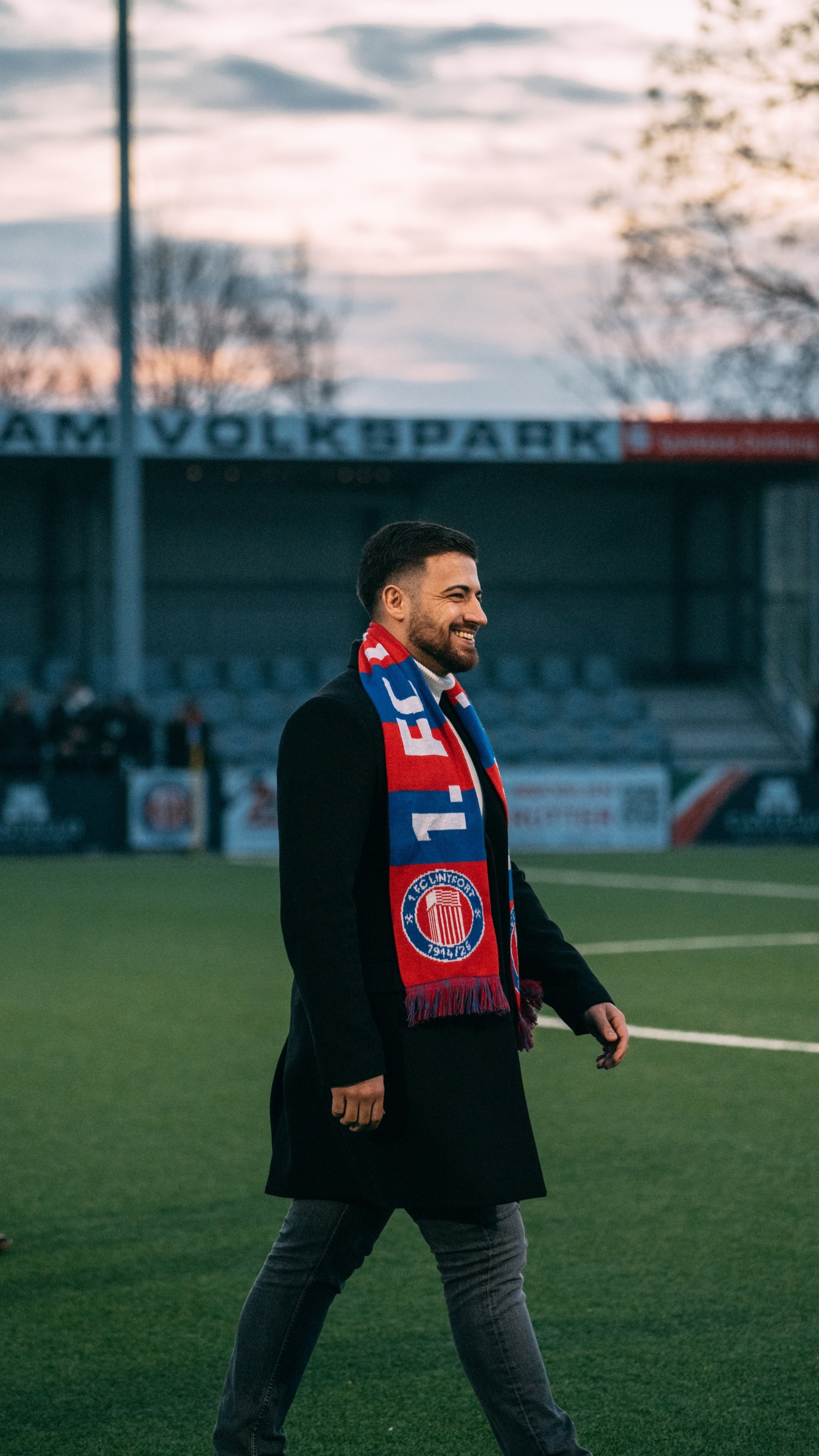 [{"clang_id":1,"value":"Mann mit Schal eines Fußballvereins geht lächelnd auf einem Fußballfeld vor einem Stadion. Der Himmel ist bewölkt, es ist Dämmerung."}]