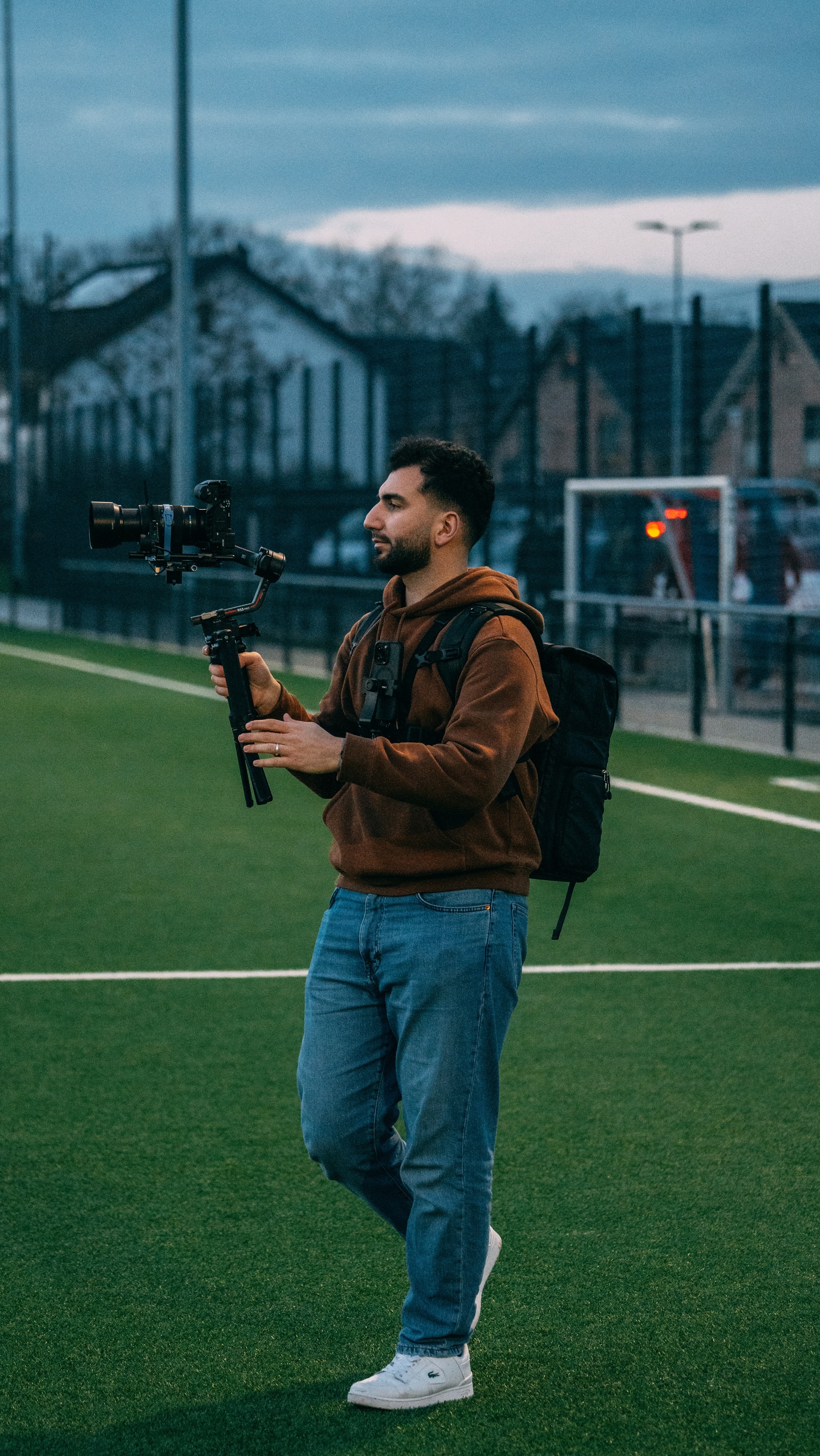 [{"clang_id":1,"value":"Mann mit Kamera auf Gimbal auf grünem Kunstrasenplatz, trägt braune Jacke, Jeans und Rucksack. Im Hintergrund Gebäude und Fußballtor."}]