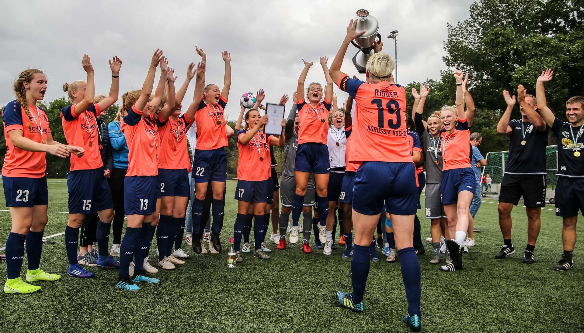[{"clang_id":1,"value":"Frauenfußballteam in orangefarbenen Trikots feiert Sieg mit Pokal. Spielerinnen heben Arme hoch, jubeln auf einem Fußballfeld. Im Hintergrund Zuschauer."}]