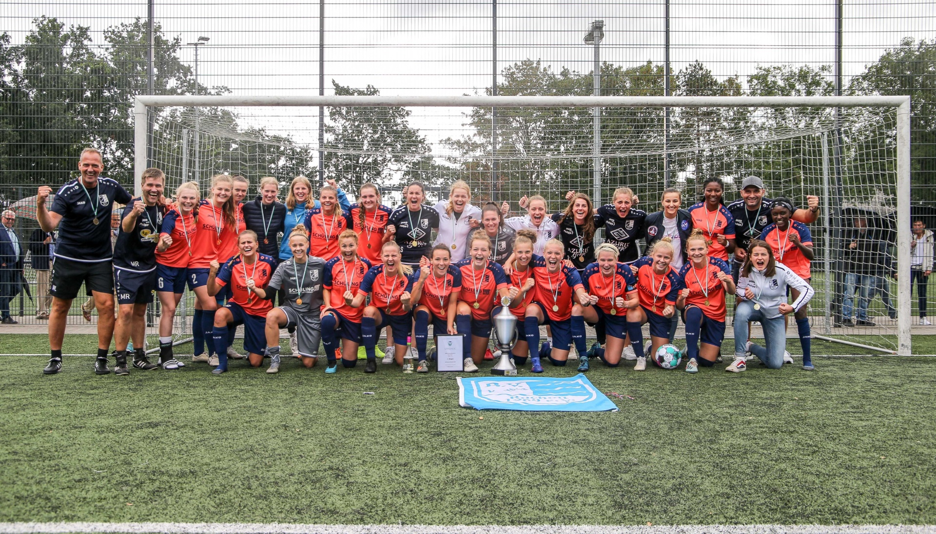 [{"clang_id":1,"value":"Fußballmannschaft mit Medaillen und Pokal auf dem Spielfeld vor einem Fußballtor. Spielerinnen und Trainer stehen oder knien."}]