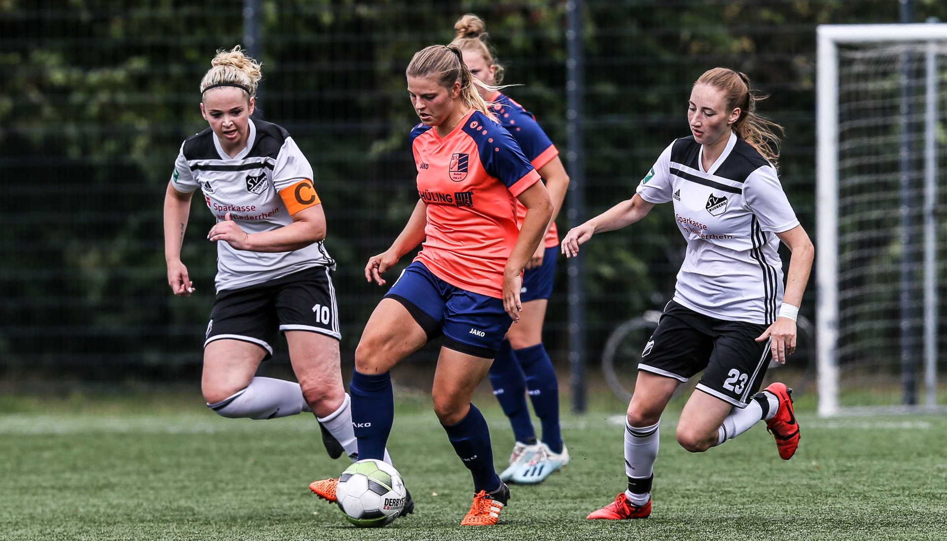 [{"clang_id":1,"value":"Drei Fußballspielerinnen auf grünem Rasen. Links in schwarz-weiß, Mitte orange, rechts schwarz-weiß. Eine Spielerin schießt auf den Ball. Im Hintergrund ein Zaun und ein Fußballtor."}]