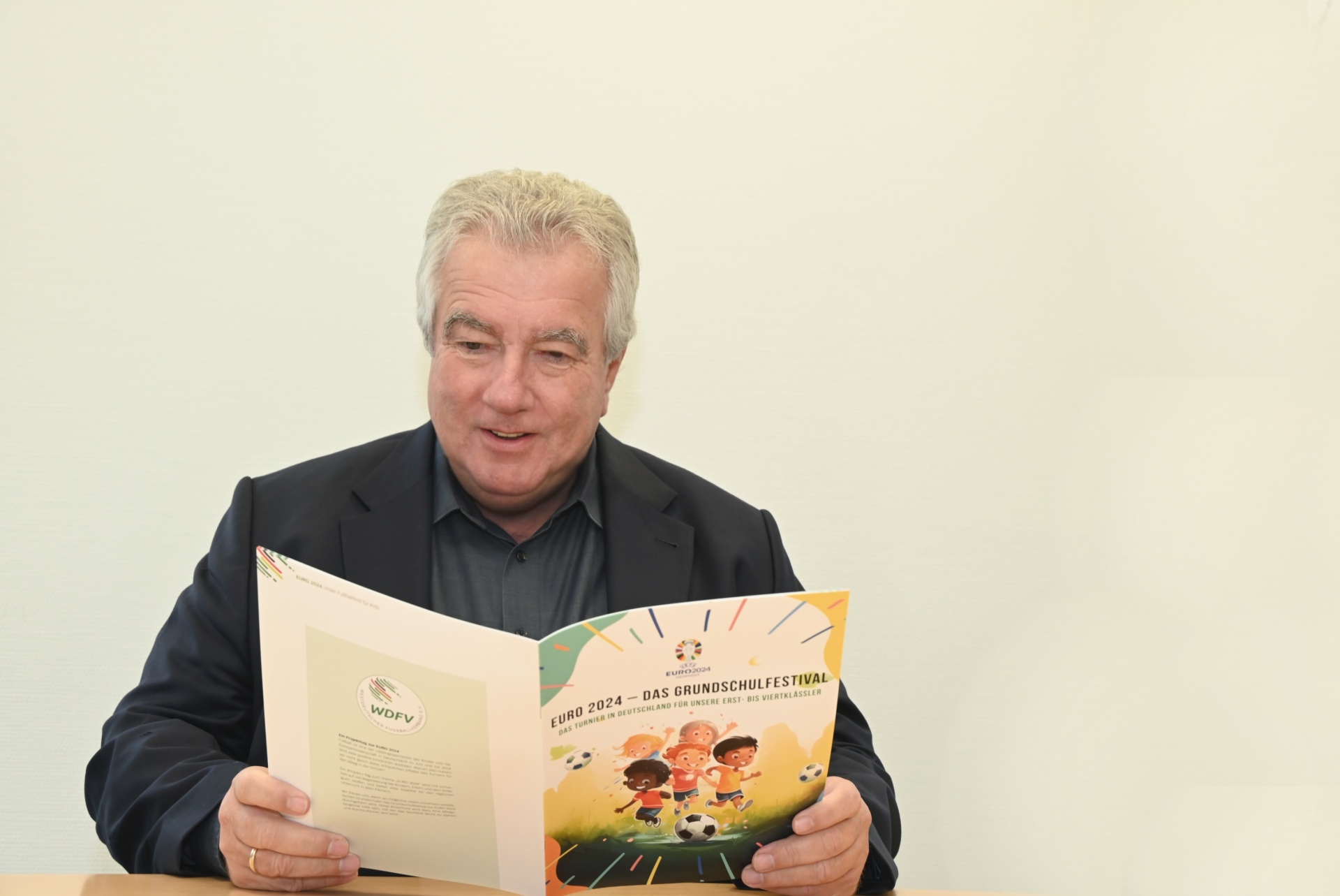 [{"clang_id":1,"value":"Mann mit grauen Haaren im schwarzen Pullover liest eine farbige Broschüre mit dem Titel \"EURO 2024 – DAS GRUNDSCHULFESTIVAL\". Hintergrund ist eine weiße Wand."}]