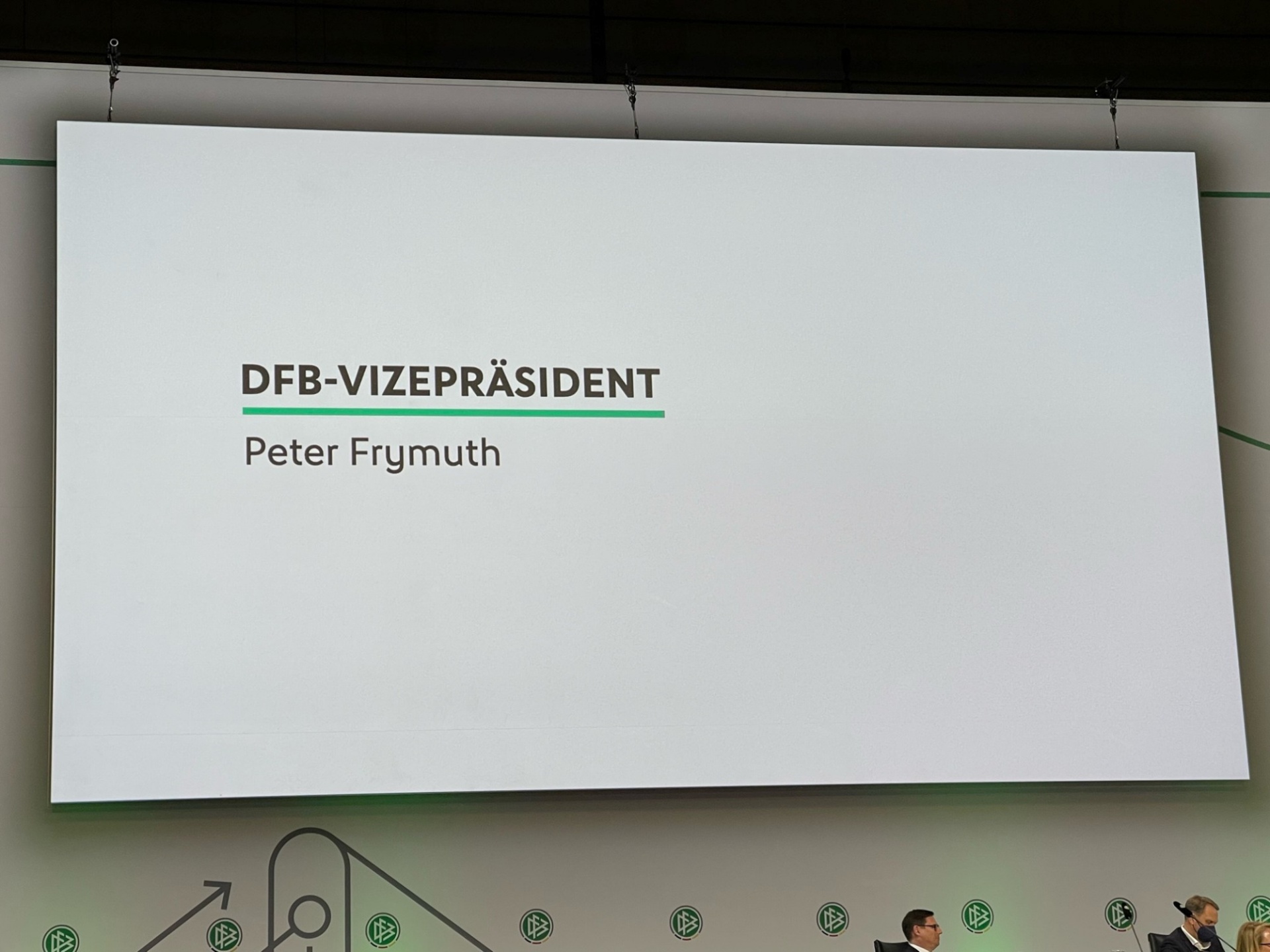 [{"clang_id":1,"value":"Präsentationsfolie: \"DFB-Vizepräsident Peter Frymuth\". Darunter eine Reihe von Personen an einem Tisch, vor grünem Hintergrund mit Logo."}]